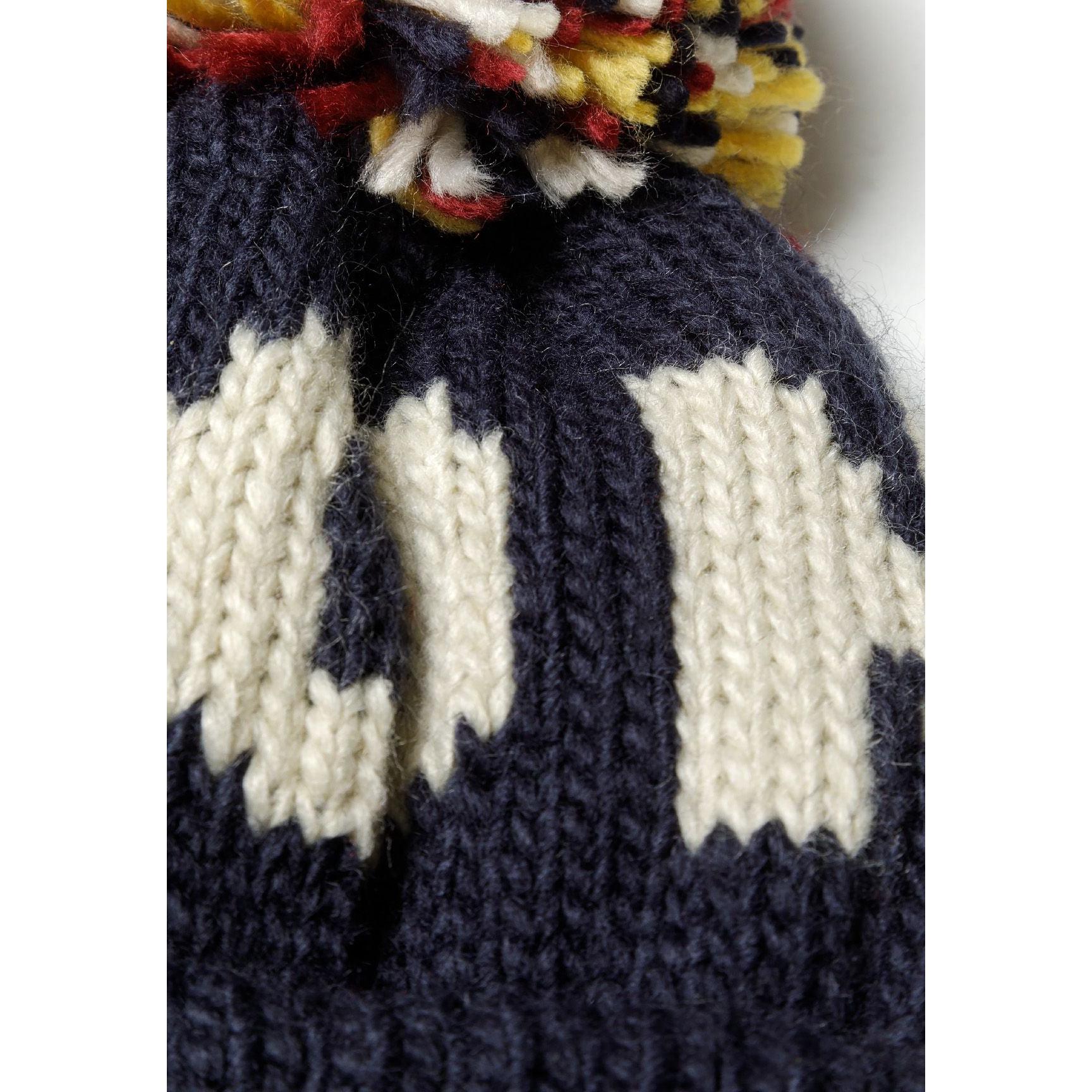 Close-up detail of navy knitted beanie showing white jacquard knit pattern and multicoloured yarn pom pom with red, yellow, white and navy strands