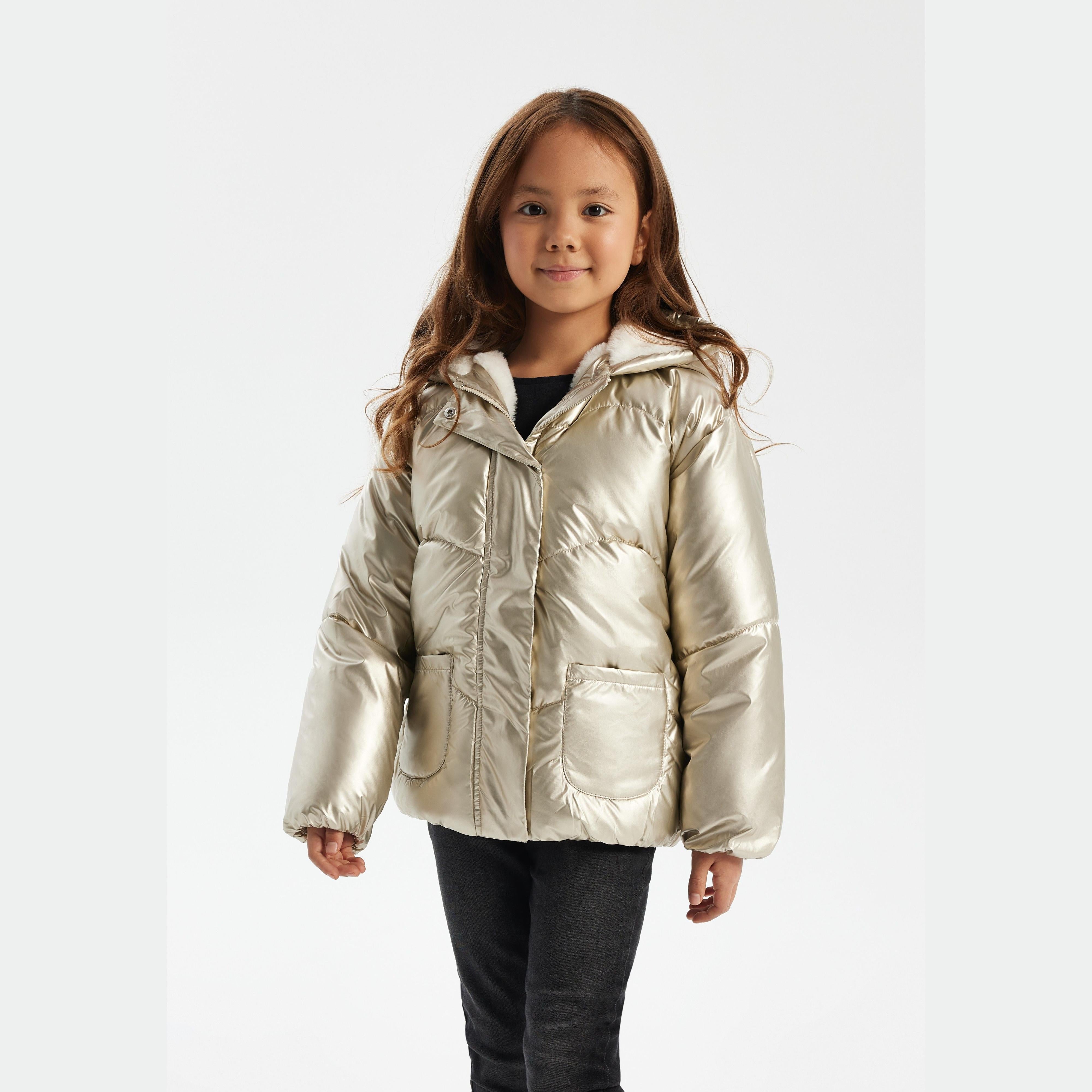 Young girl wearing shiny metallic gold padded jacket with hood up, standing against white background, paired with dark jeans
