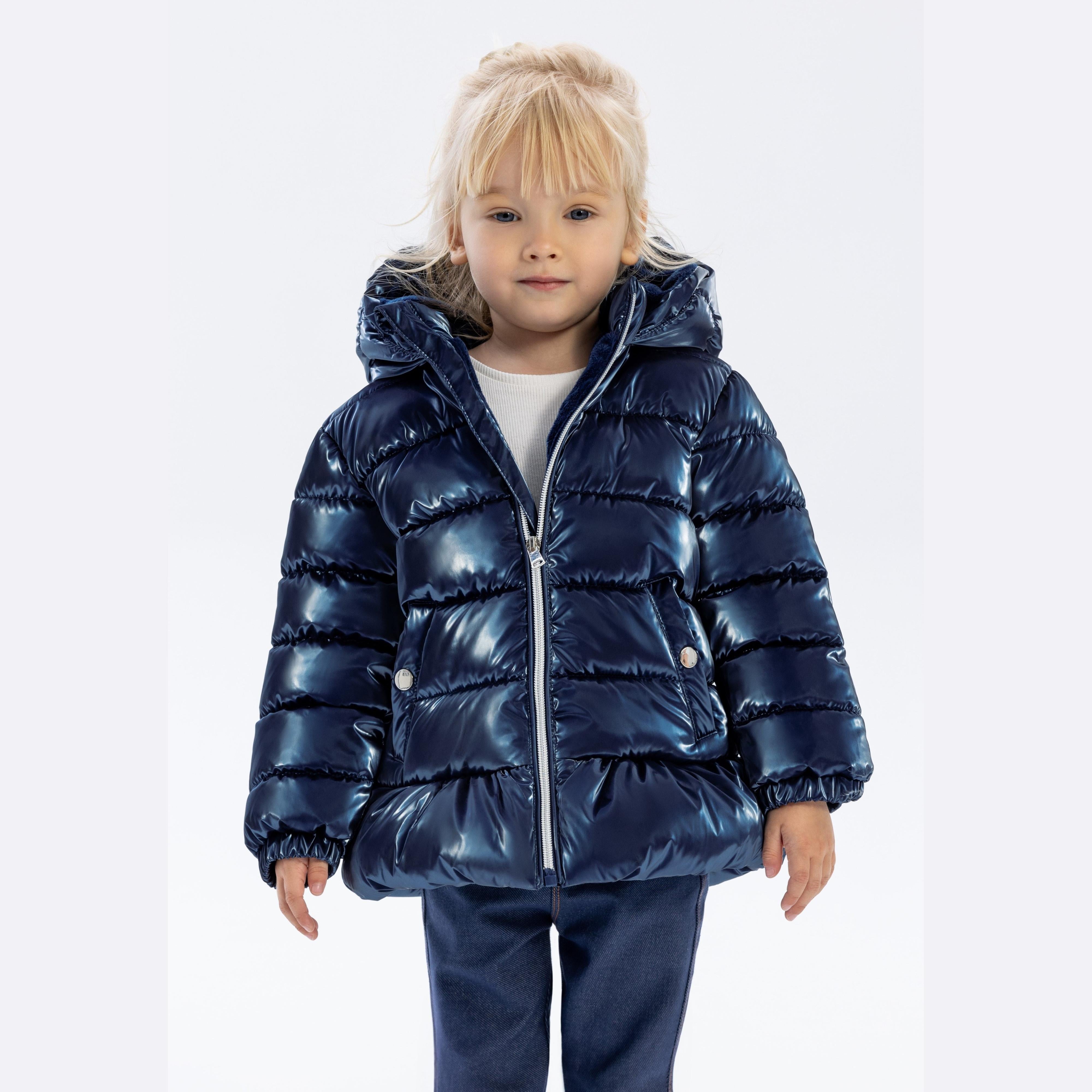 Young blonde child wearing metallic navy blue padded jacket with hood, white top underneath, standing against white background