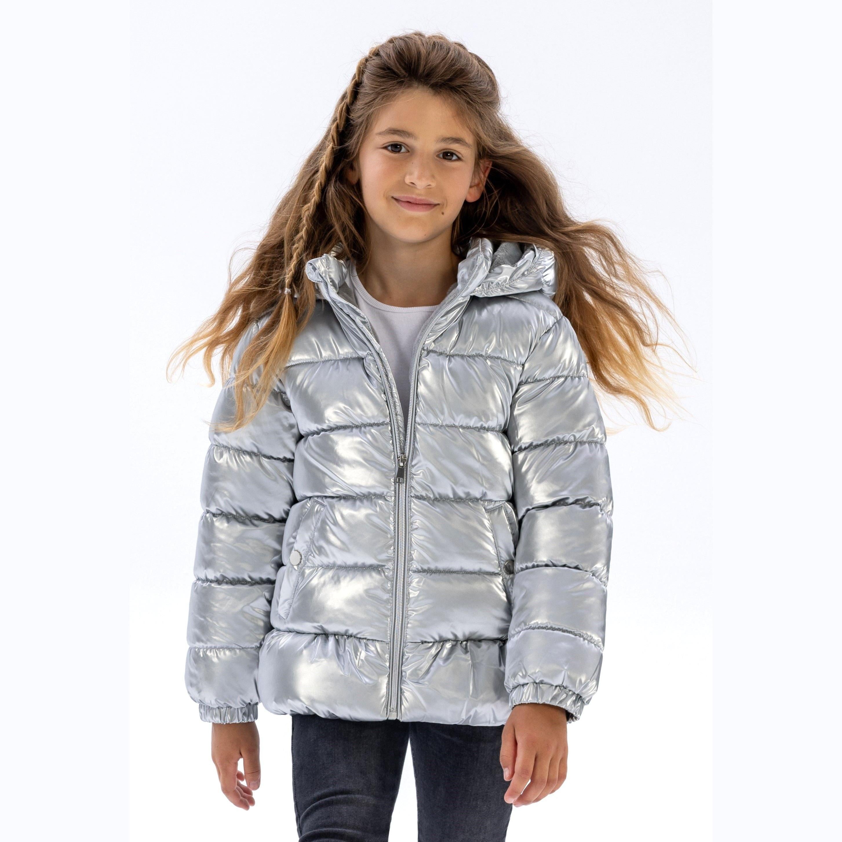 Young girl wearing silver metallic padded jacket with hood up, standing against white background, paired with dark jeans