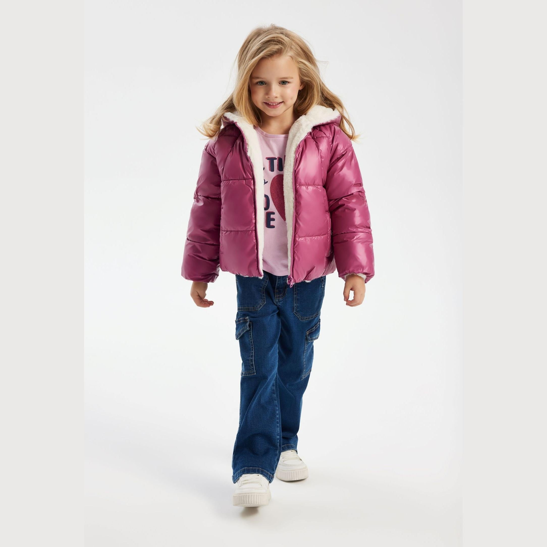 Young blonde girl wearing metallic purple reversible padded jacket with white teddy fur hood, pink top with text, blue jeans, walking on white studio background