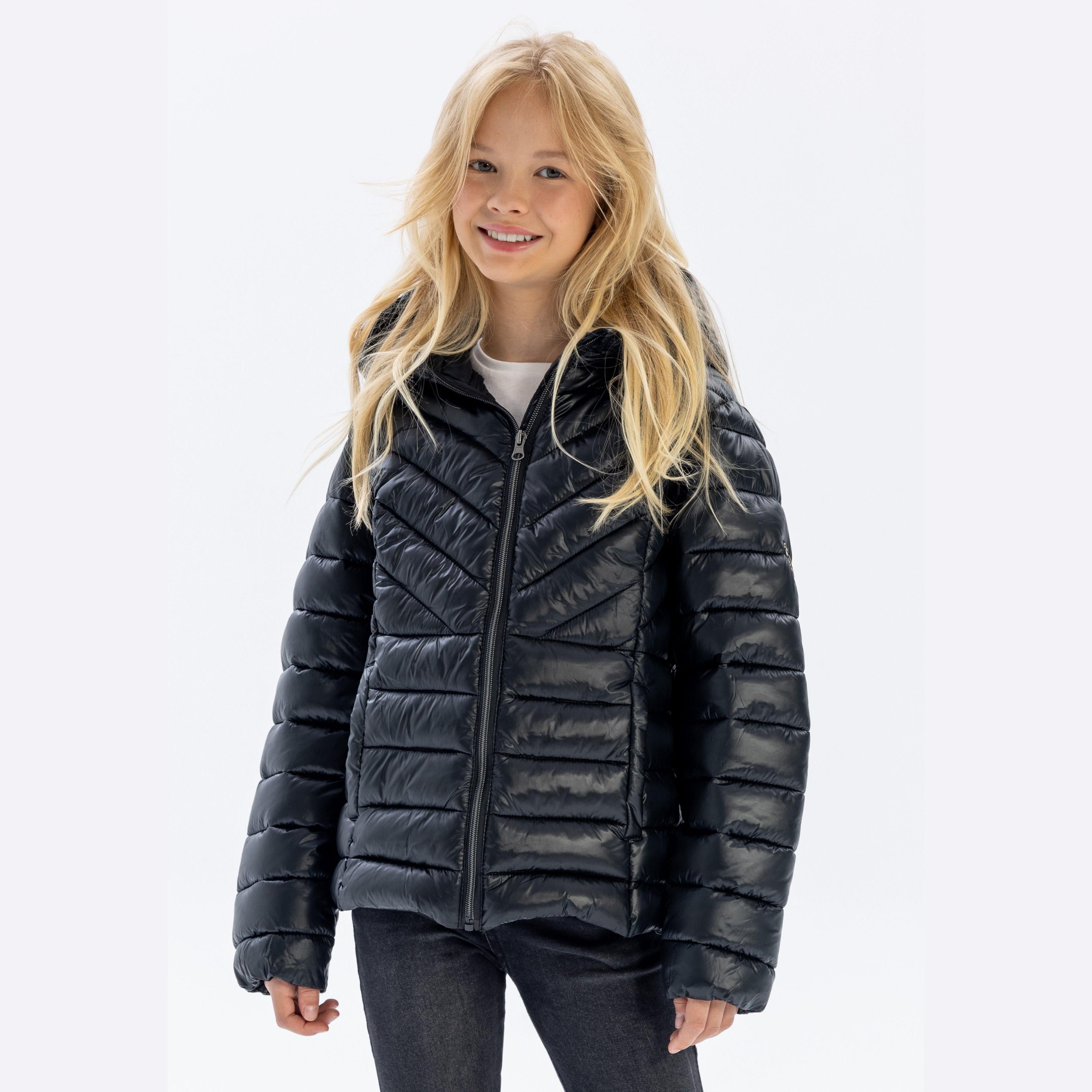 Young blonde girl wearing black quilted hooded jacket over white top with dark jeans, smiling at camera against white studio background