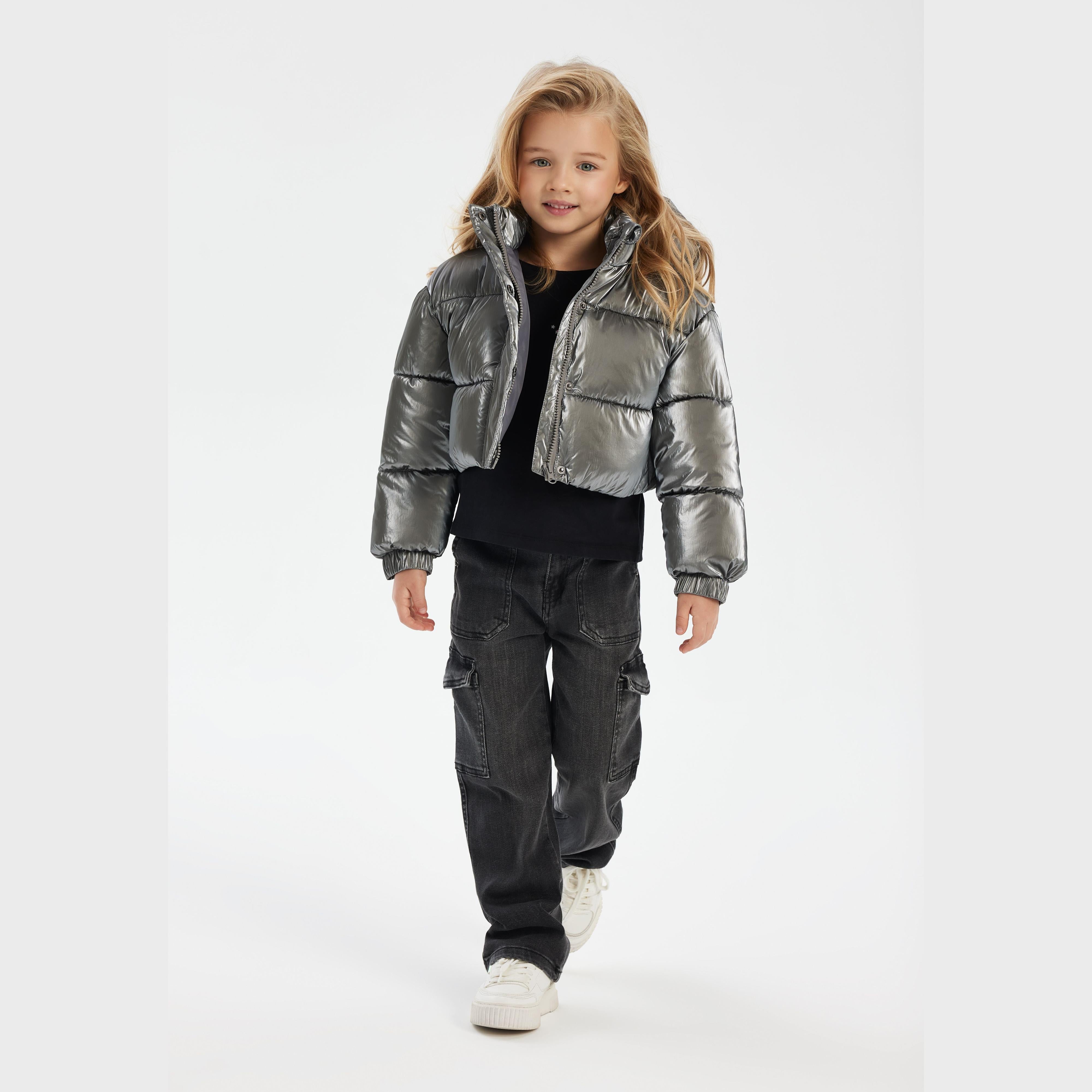 Young girl wearing grey metallic cropped padded jacket with hood up, paired with black cargo pants and white sneakers, walking against white background