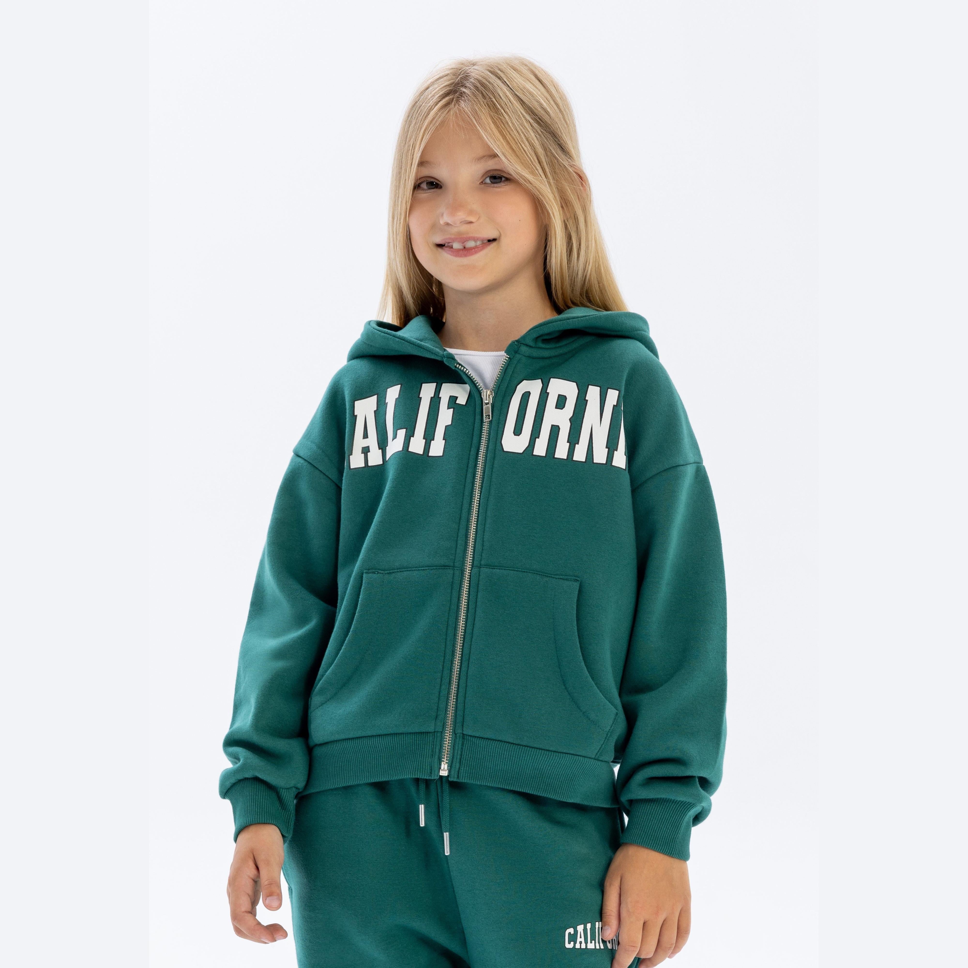 Young blonde girl wearing green California print zip hoodie with matching green tracksuit bottoms, smiling at camera against white studio background