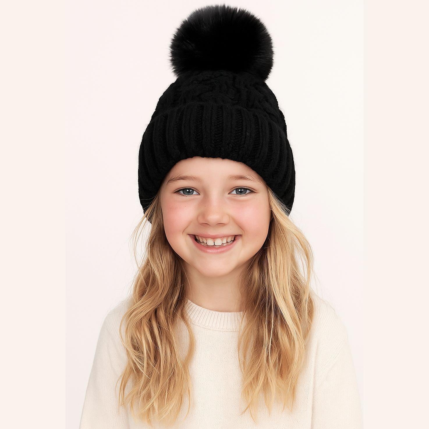 Young blonde girl wearing black cable knit hat with fluffy pom pom and cream sweater, smiling against pink background
