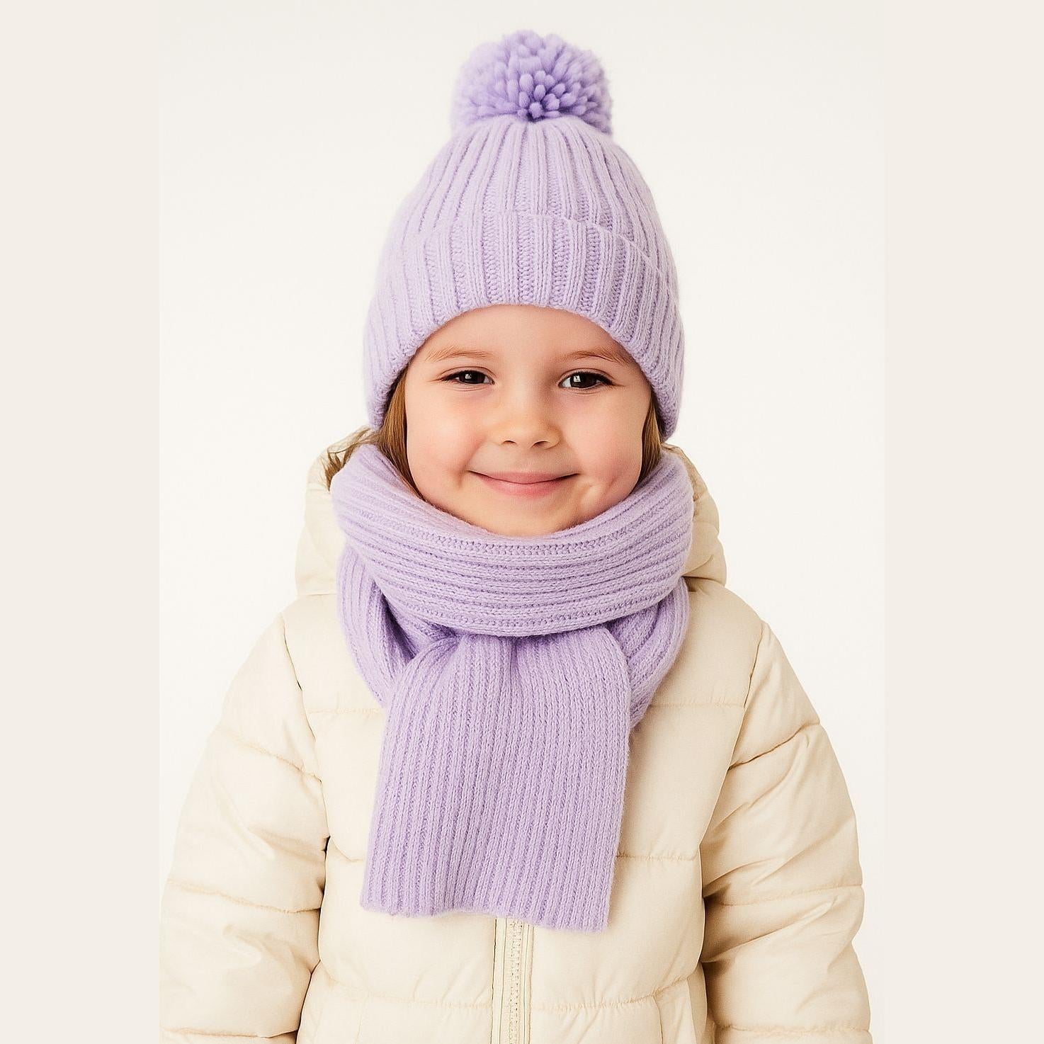 Young child wearing lilac ribbed knit winter hat with pom pom and matching scarf, cream jacket, studio white background