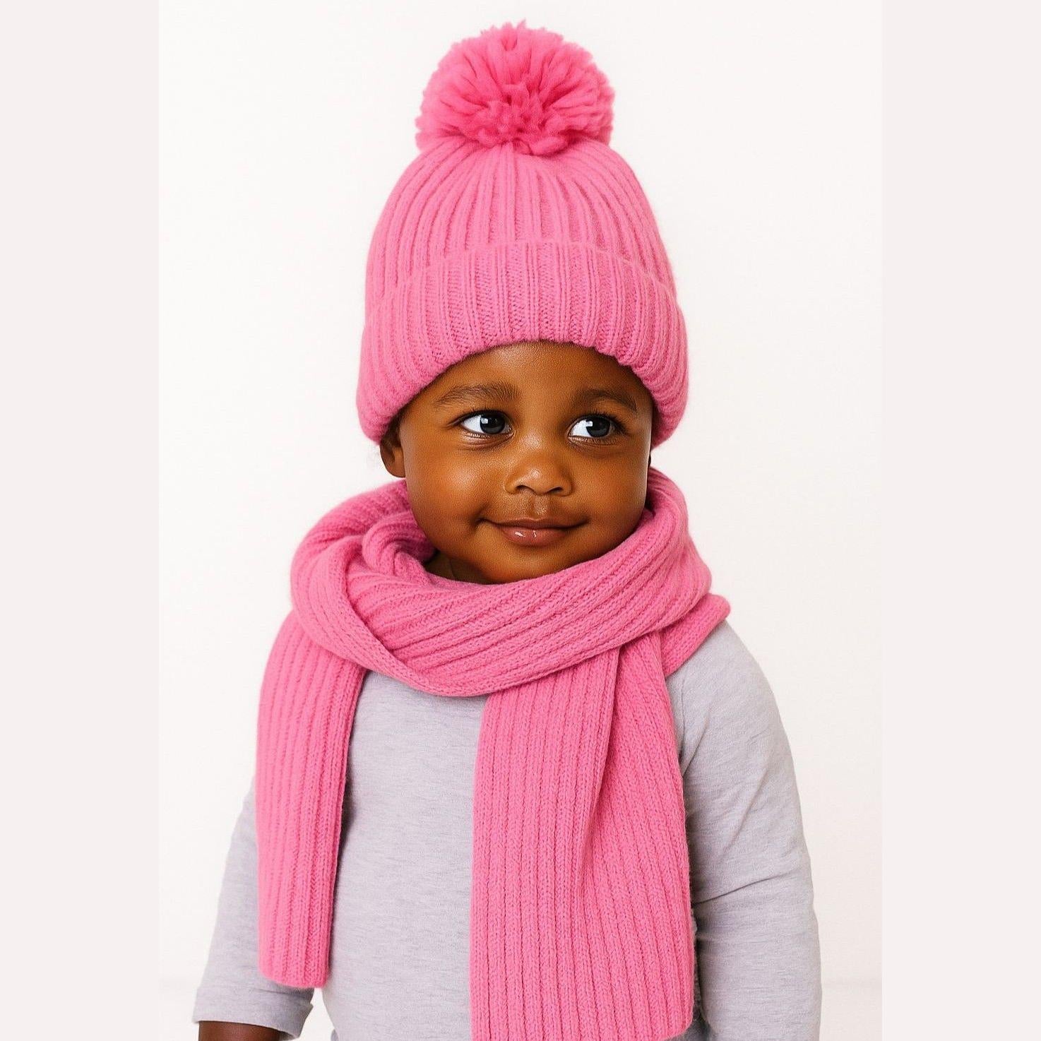 Young child wearing pink ribbed knit beanie hat with pom pom and matching scarf over grey top against white background