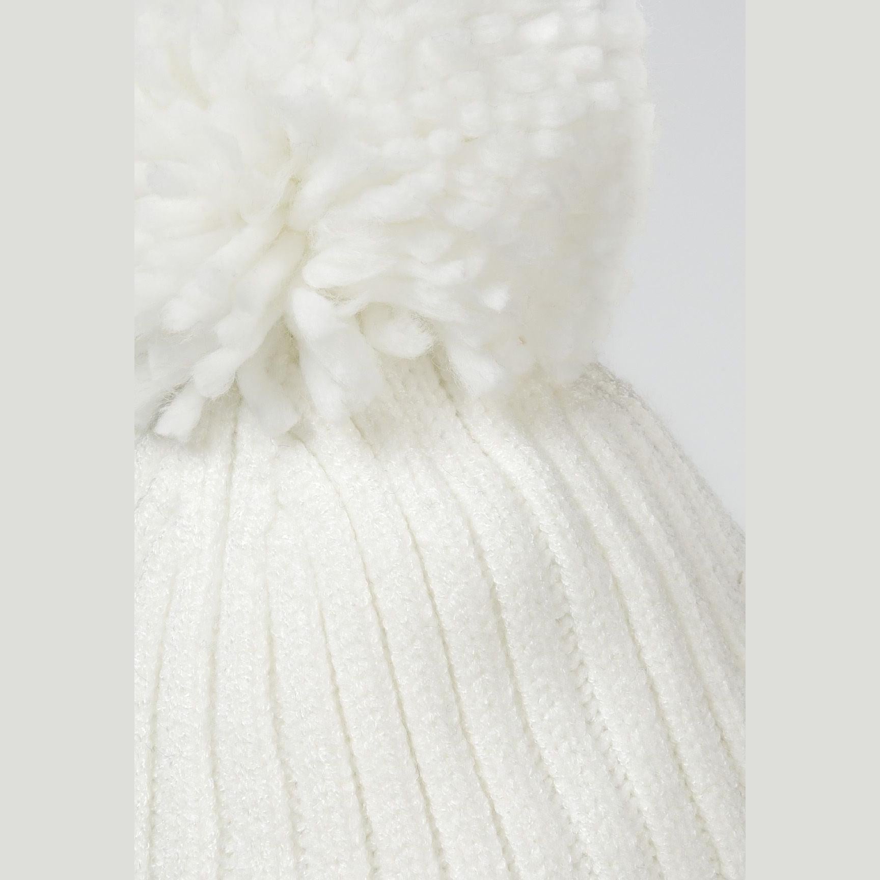 Close-up detail of cream knitted hat showing ribbed texture and pom pom attachment point at crown