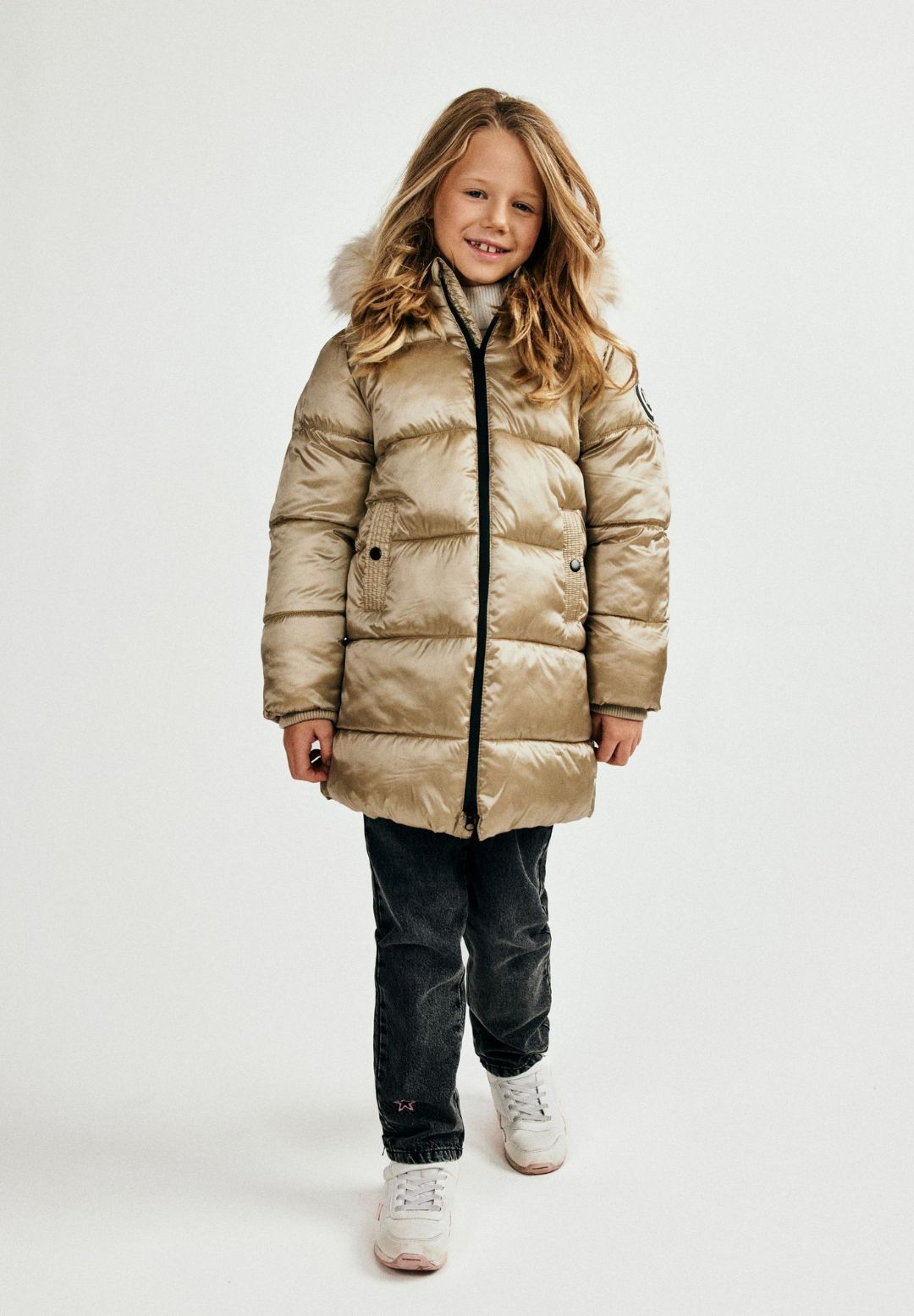 Girl wearing a champagne-gold quilted parka with black front zip, snap-tab pockets and fur-look hood trim.