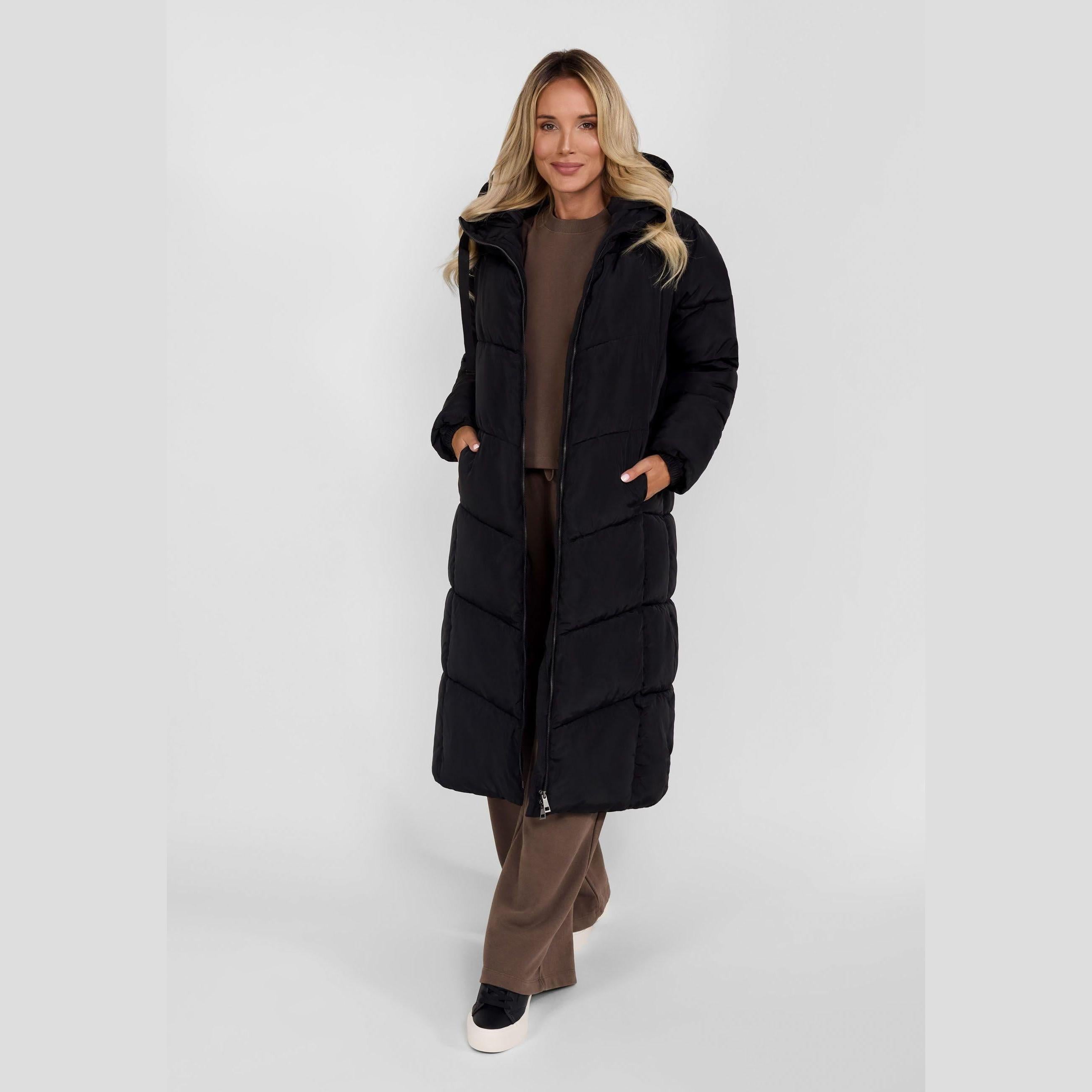 Woman wearing long black quilted winter coat with hood over brown outfit against white background