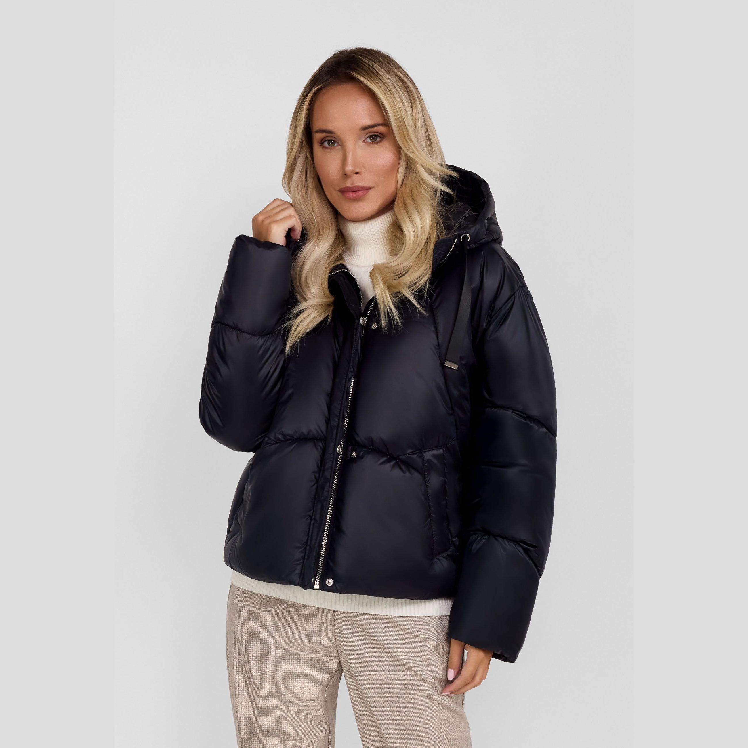 Woman wearing black quilted puffer jacket with hood, cream turtleneck sweater and beige trousers against white studio background