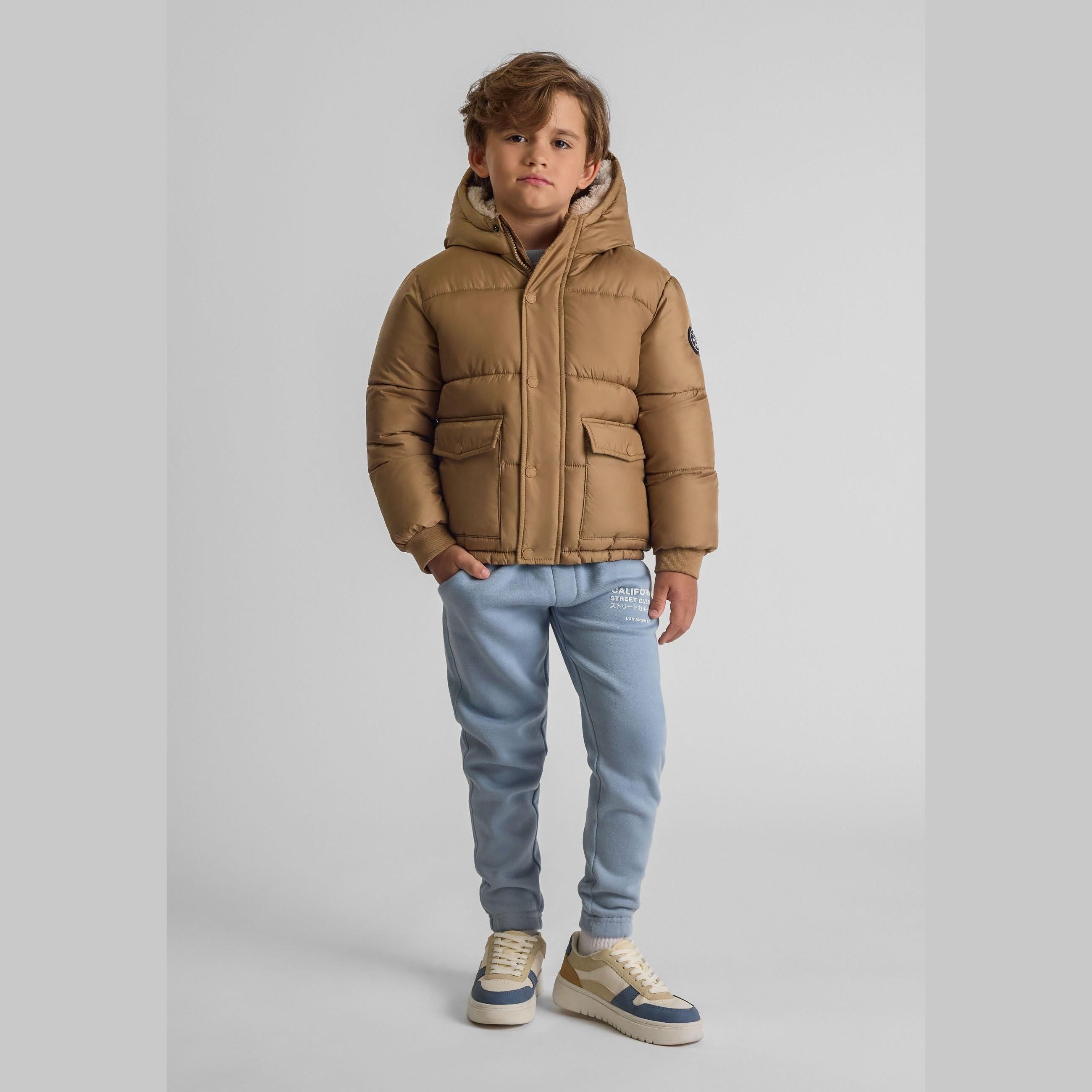 Boy wearing beige padded jacket with hood up, light blue jeans and sneakers, standing against white background