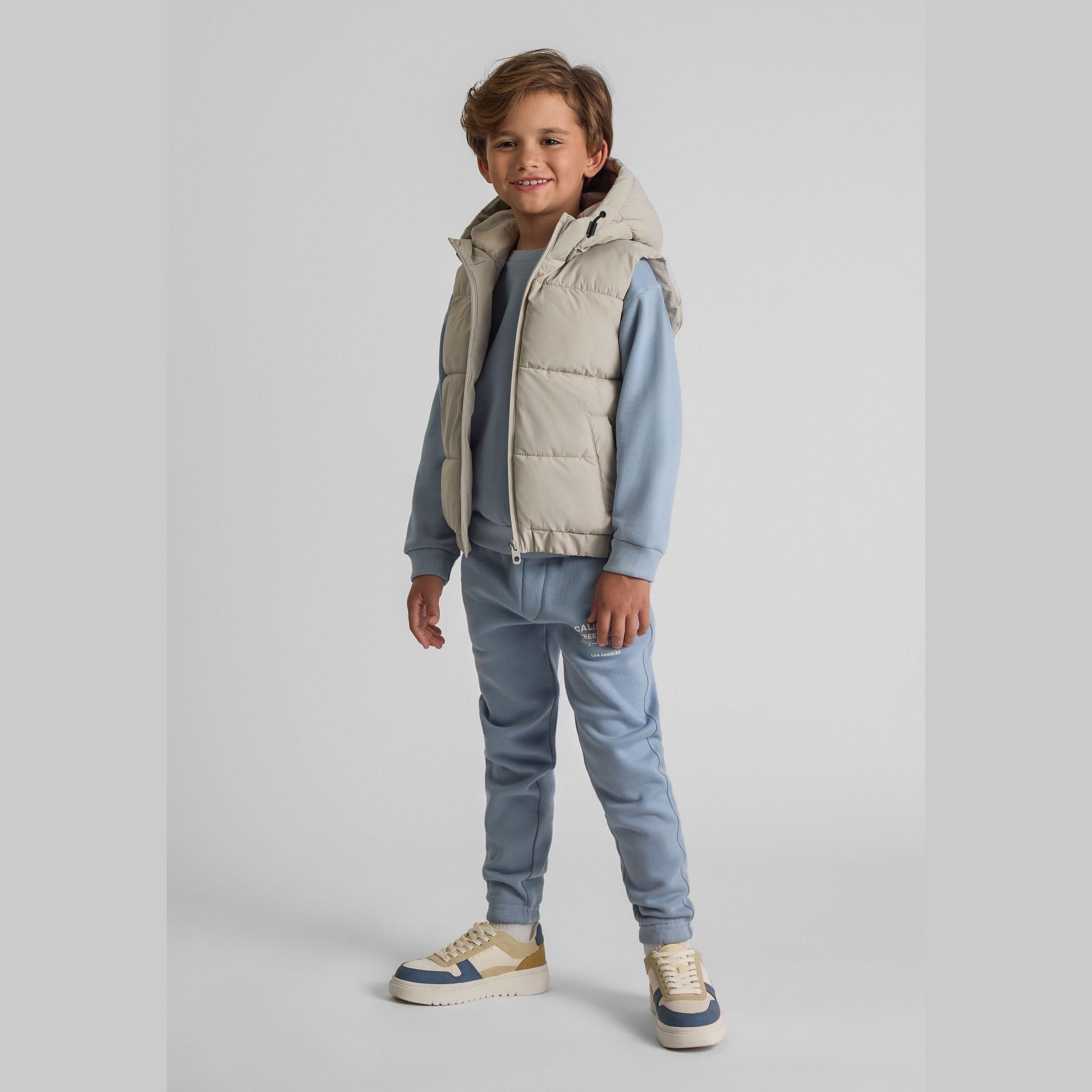 Young boy wearing grey padded ripstop gilet with hood over blue hoodie and matching blue joggers, standing on white background