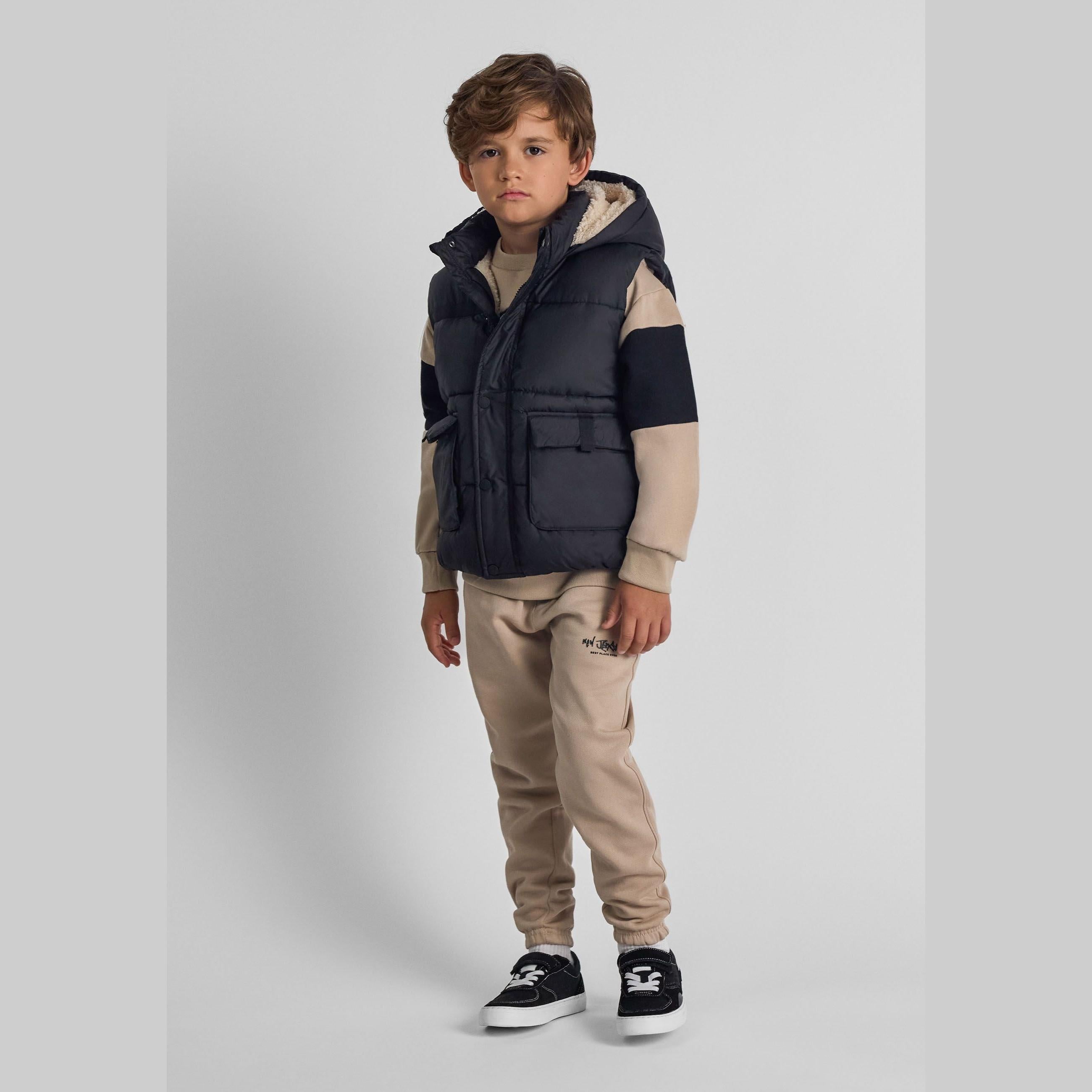 Young boy wearing black hooded gilet over beige striped sweater with beige joggers and black sneakers, standing against white background