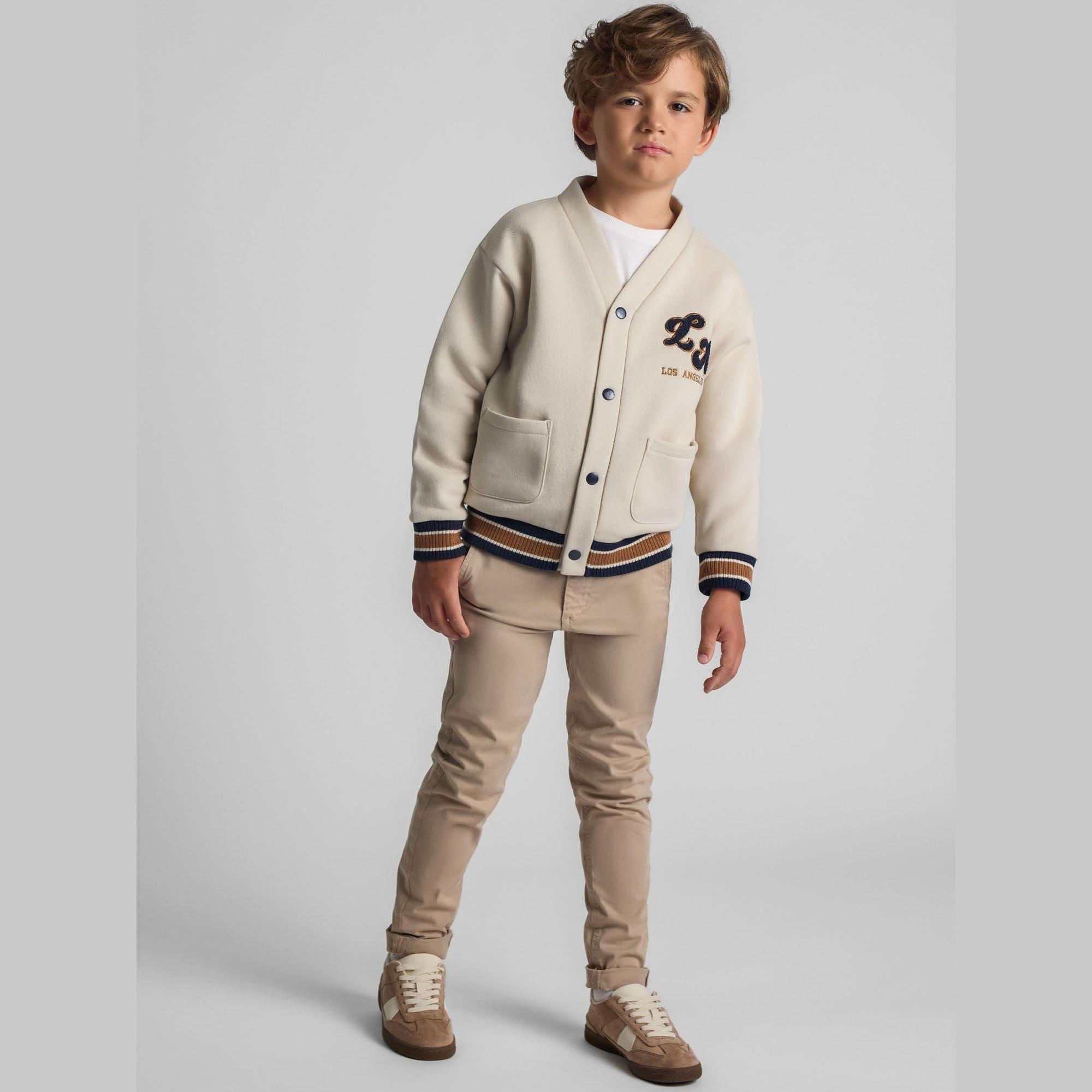 Boy wearing cream cardigan with embroidered chest applique, button-front closure and varsity-style ribbed trim, paired with beige trousers against light background