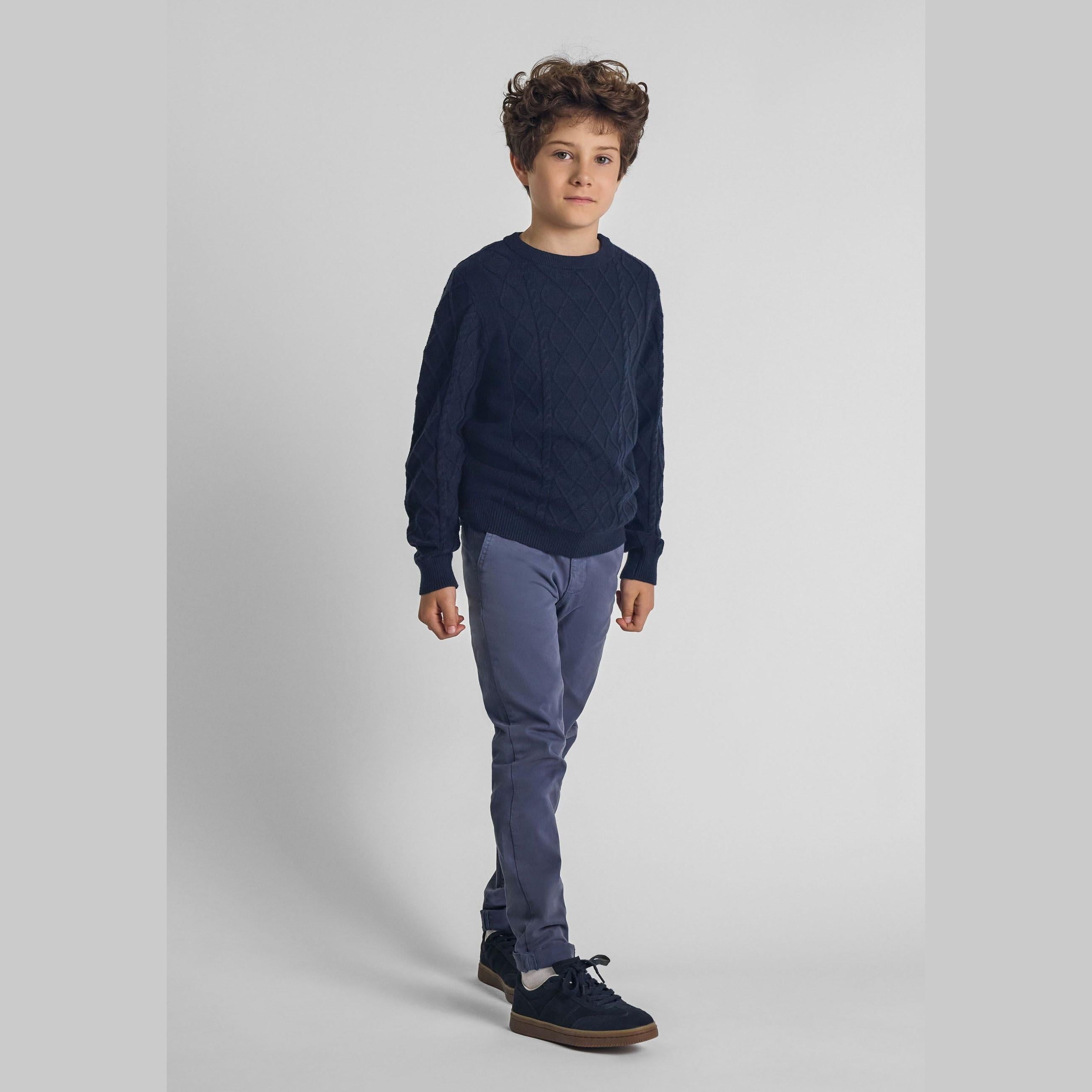 Boy wearing navy cable knit crew neck jumper with light blue trousers and dark sneakers, standing against white studio background