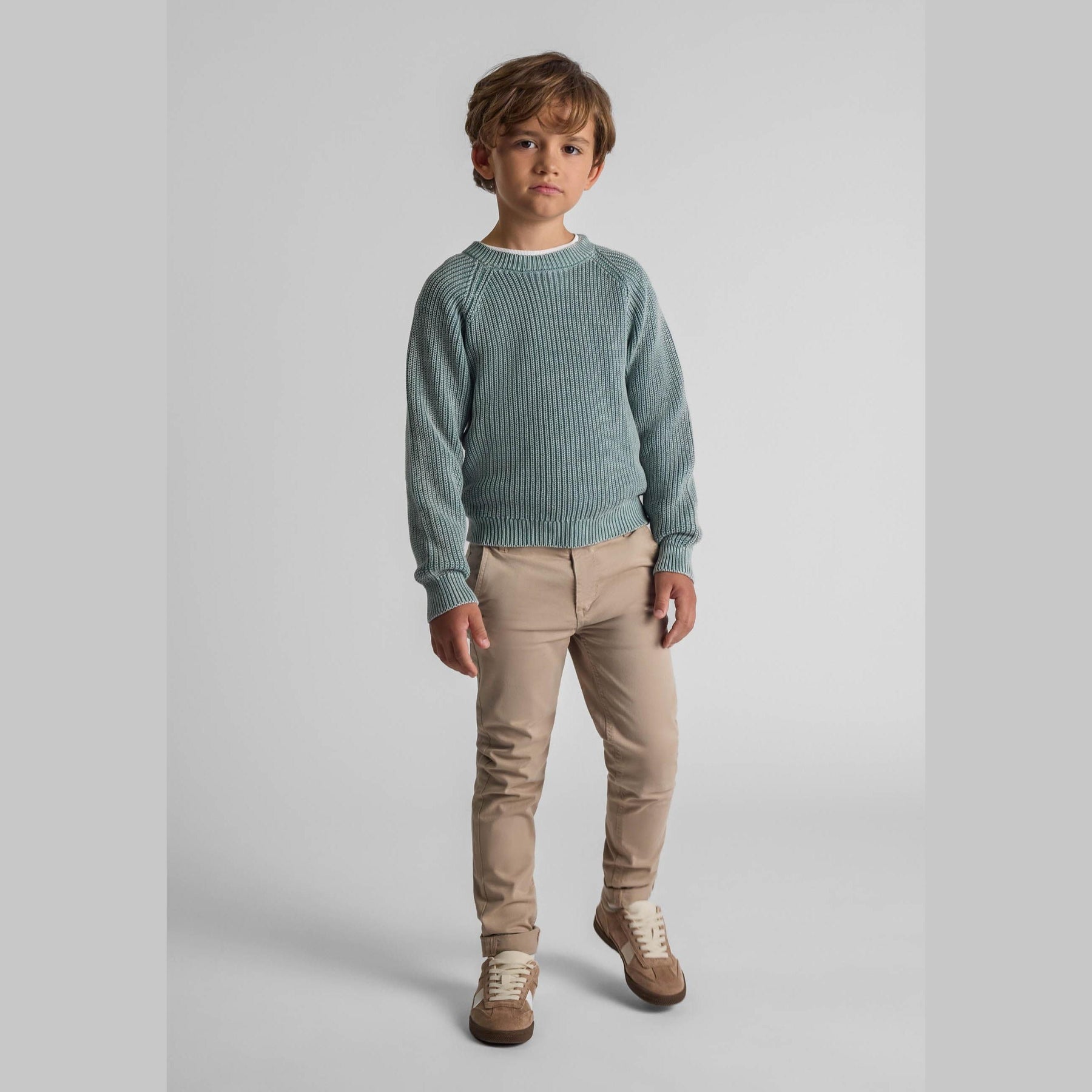 Young boy wearing khaki knit jumper with raglan sleeves and beige trousers, standing against light grey background in studio setting