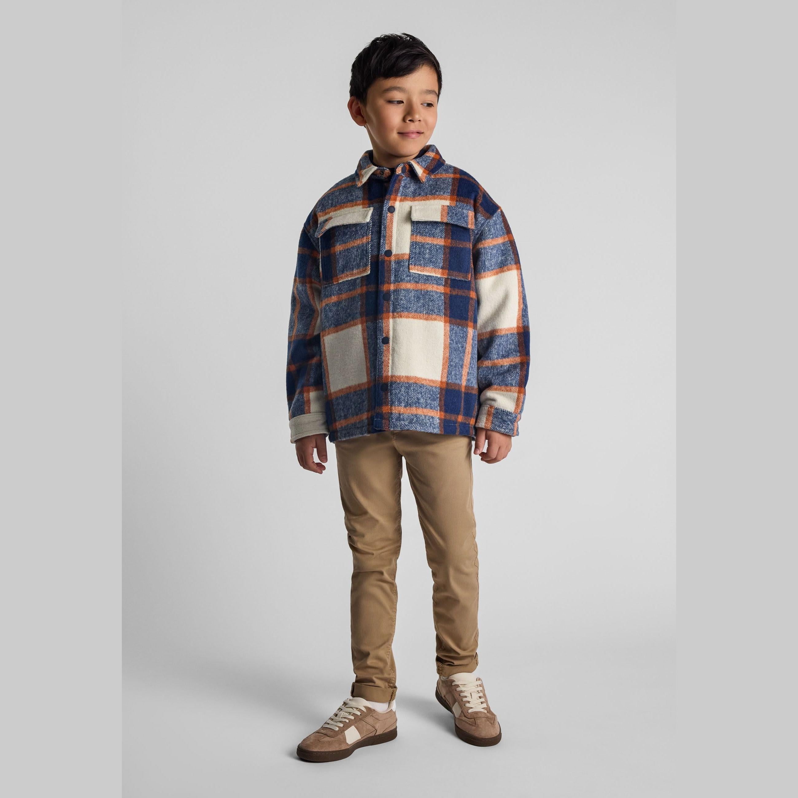 Young boy wearing blue check padded shirt jacket with beige trousers and brown sneakers against grey studio background