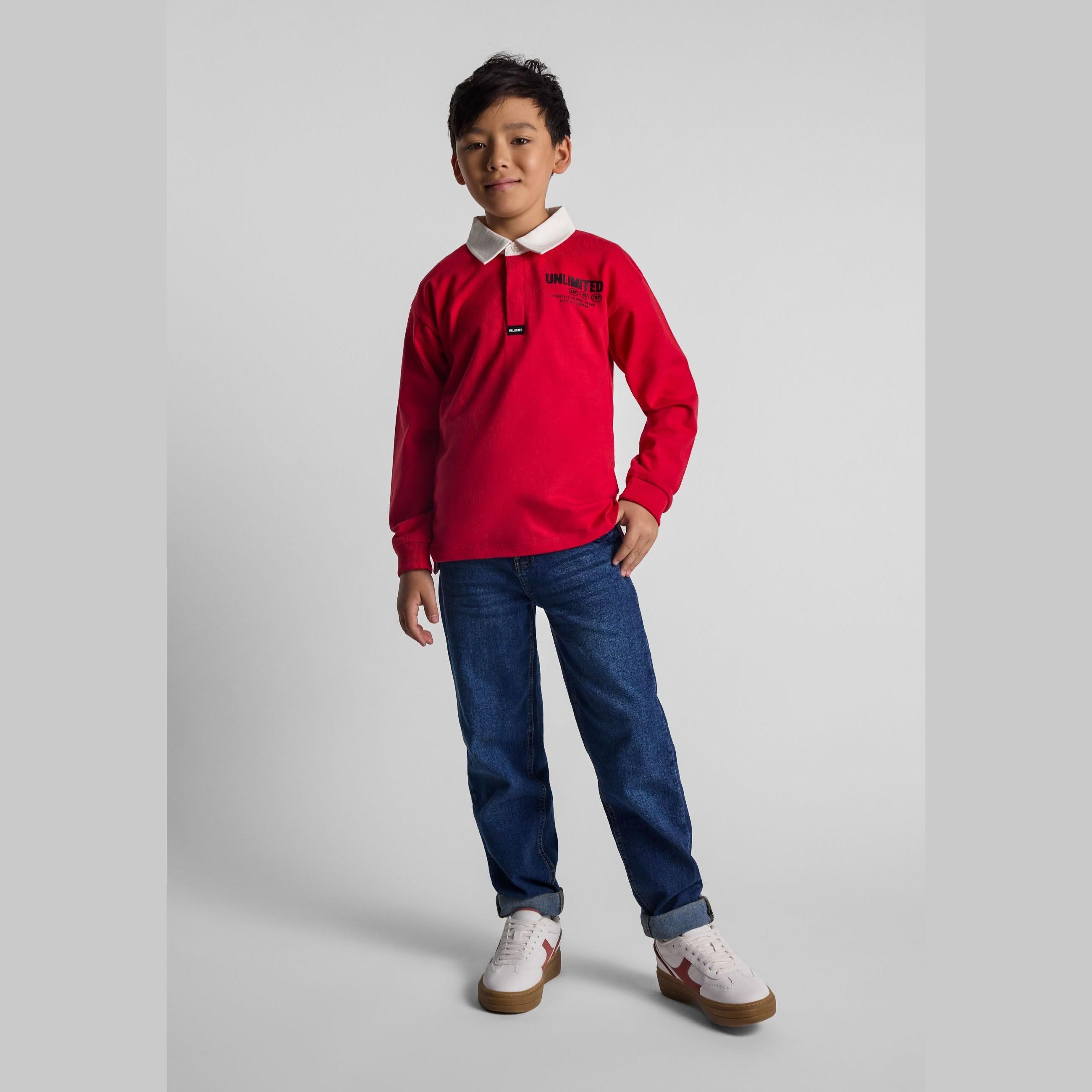 Young boy wearing red long sleeve polo shirt with white collar and dark jeans, standing against light grey background in studio setting