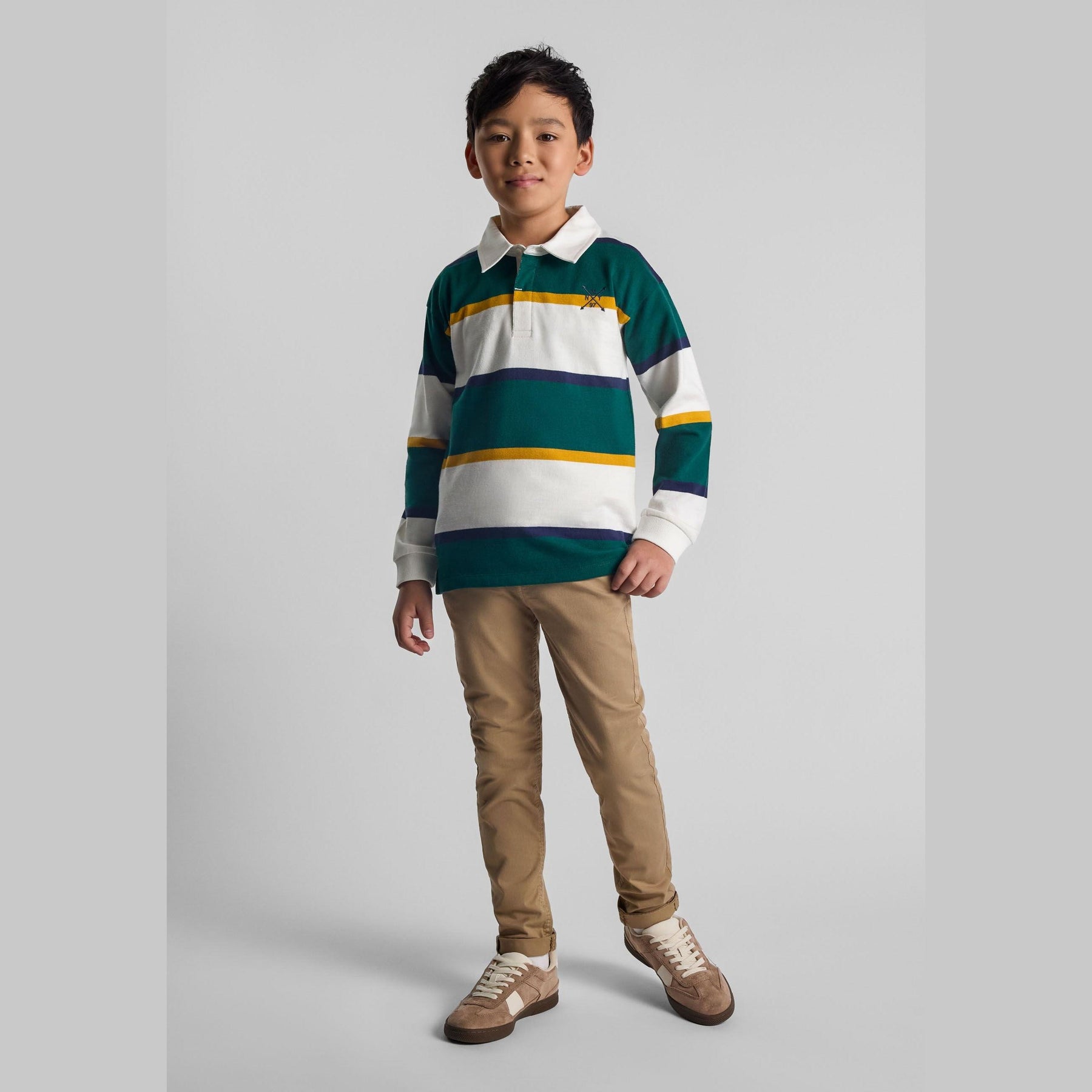 Young boy wearing green and white striped long sleeve polo shirt with khaki trousers and sneakers, standing against white background