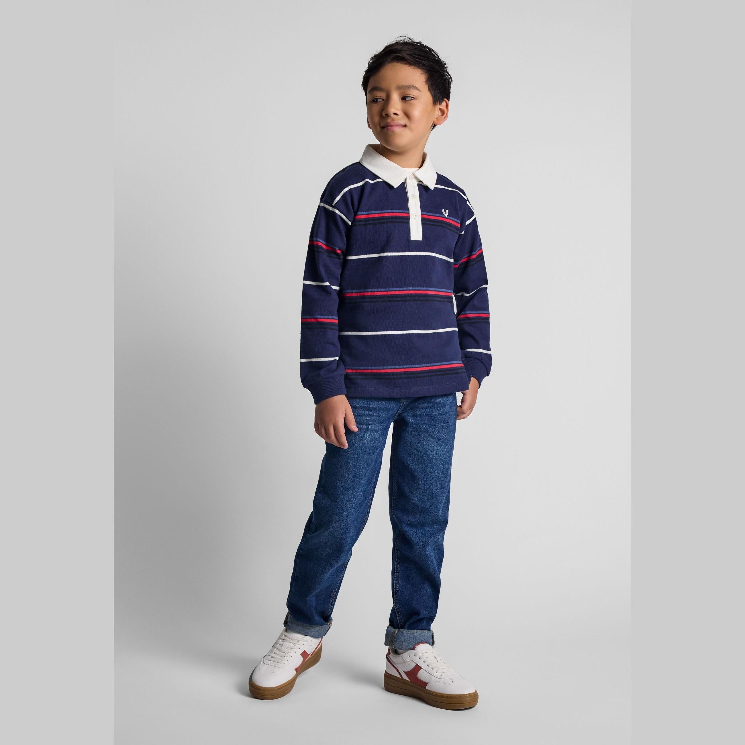Young boy wearing navy striped long sleeve polo shirt with blue jeans and white trainers, standing against neutral grey background
