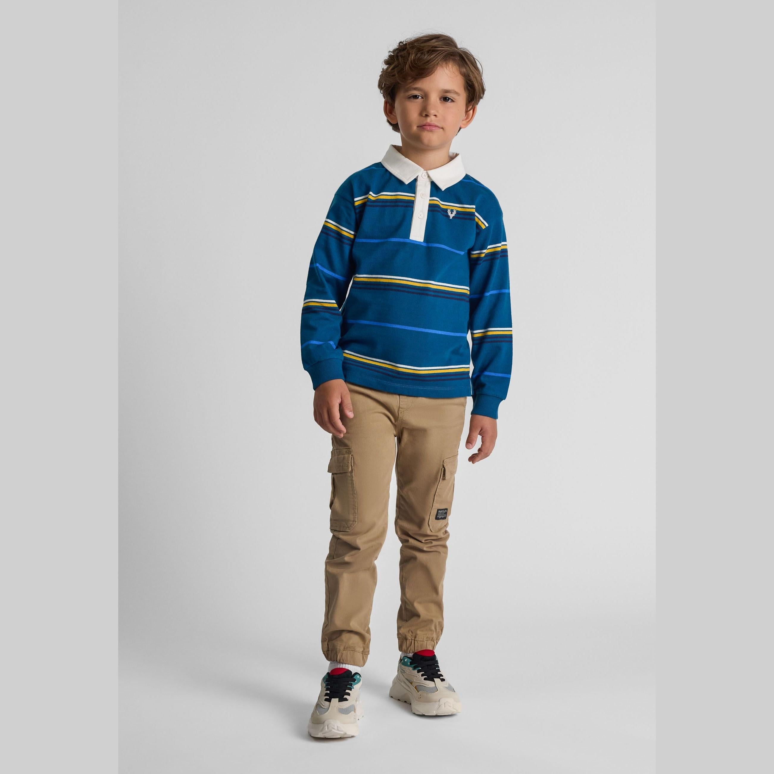Young boy wearing turquoise striped long sleeve polo shirt with white collar and beige cargo pants, standing against grey studio background