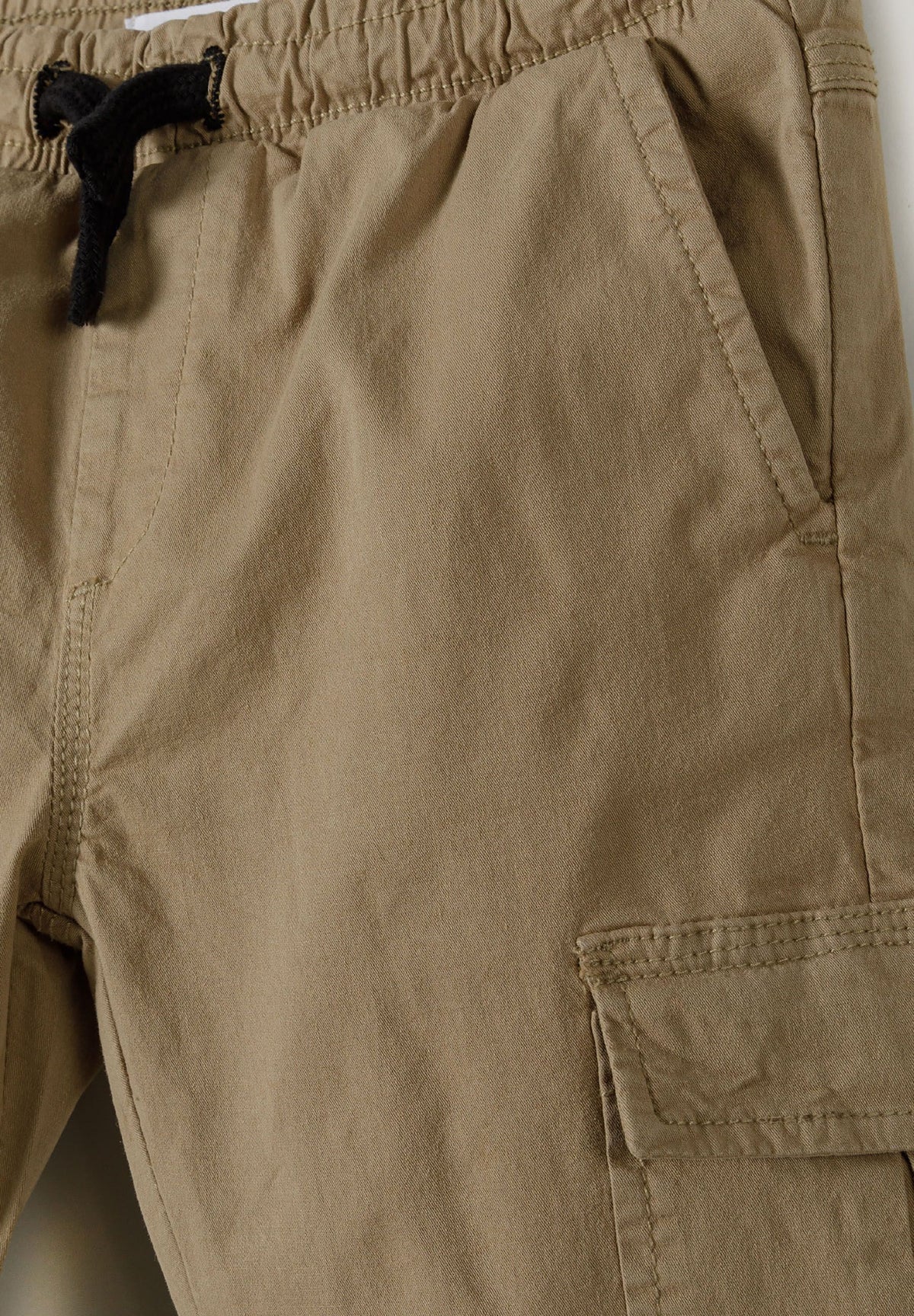 Close-up of beige cargo trousers' black drawcord, slanted hip pocket and thigh flap.