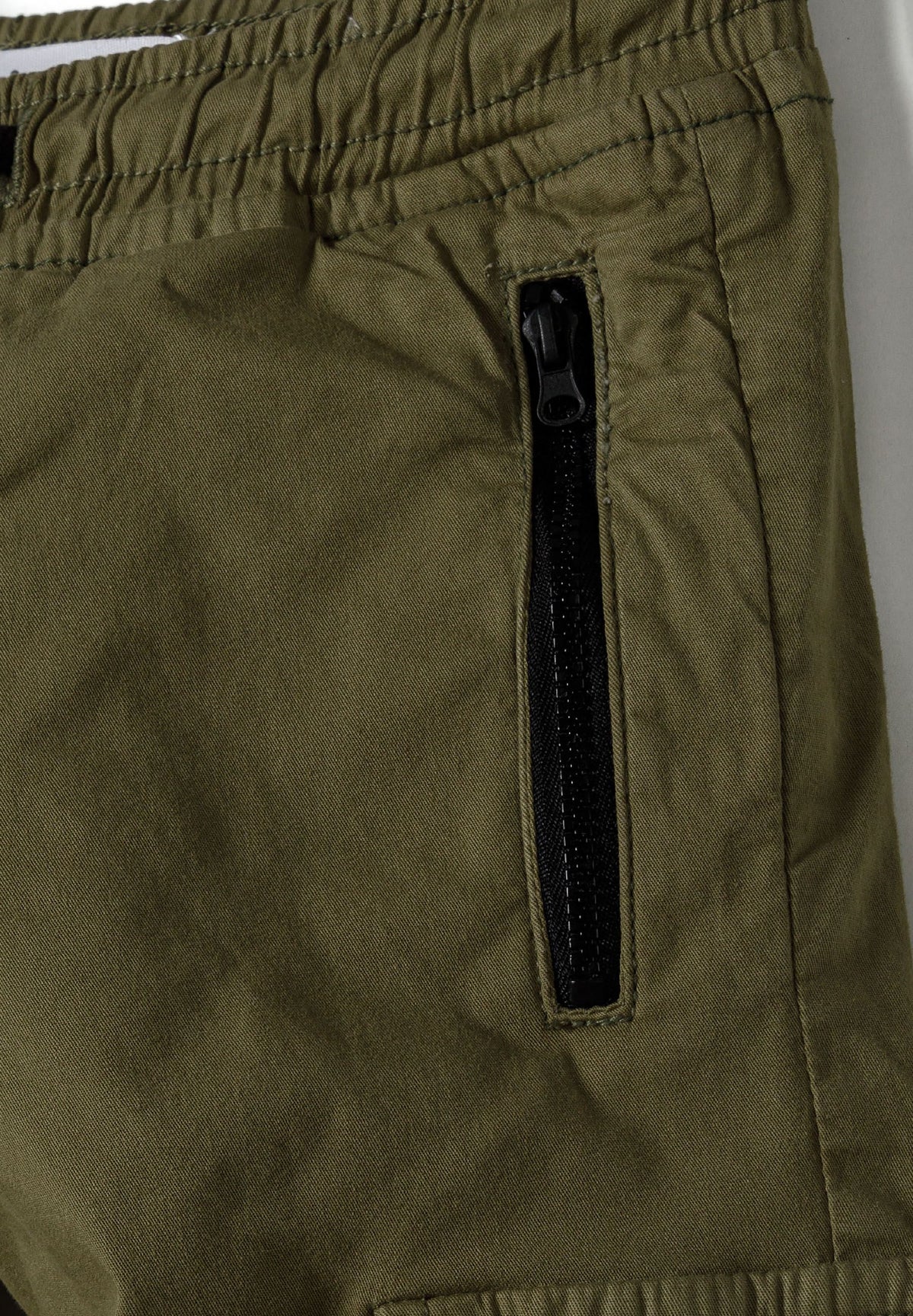Close-up of black vertical zip pocket beside gathered waistband on olive khaki trousers.