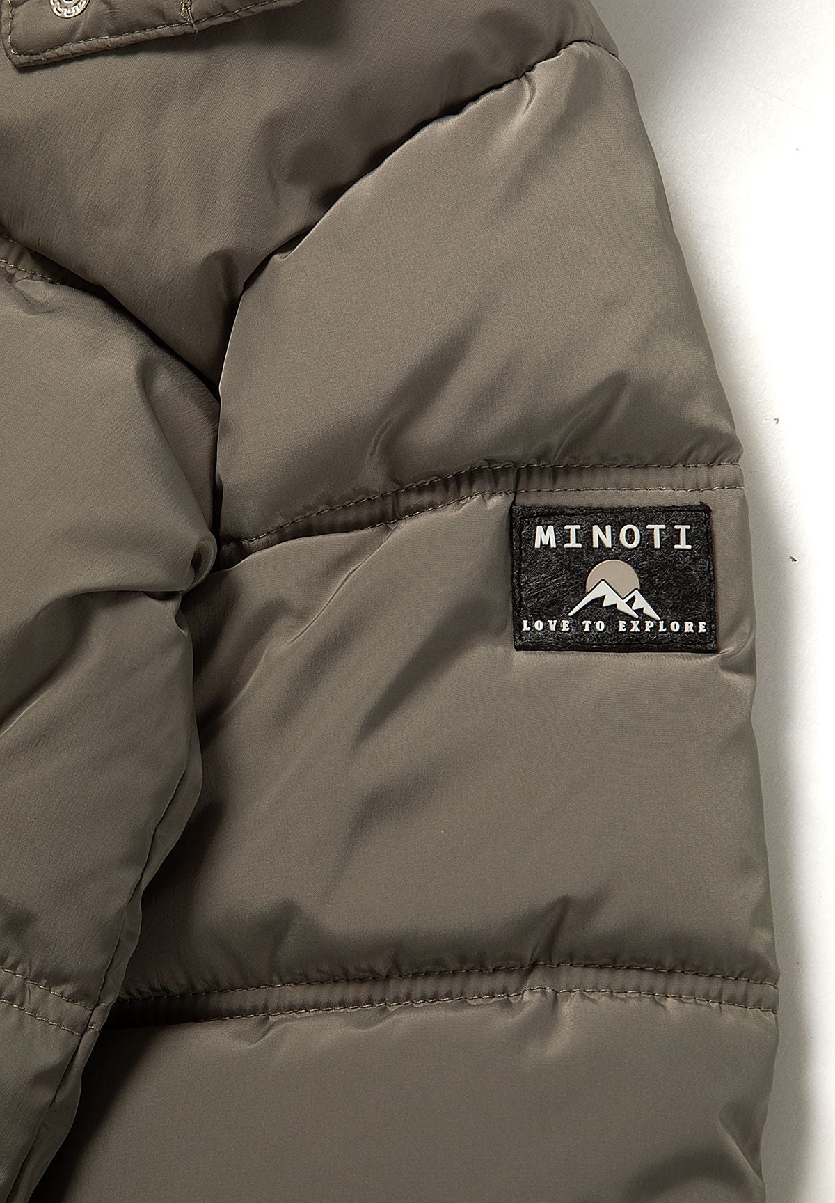 Close-up of the black Minoti mountain logo patch on the upper sleeve of the khaki jacket.