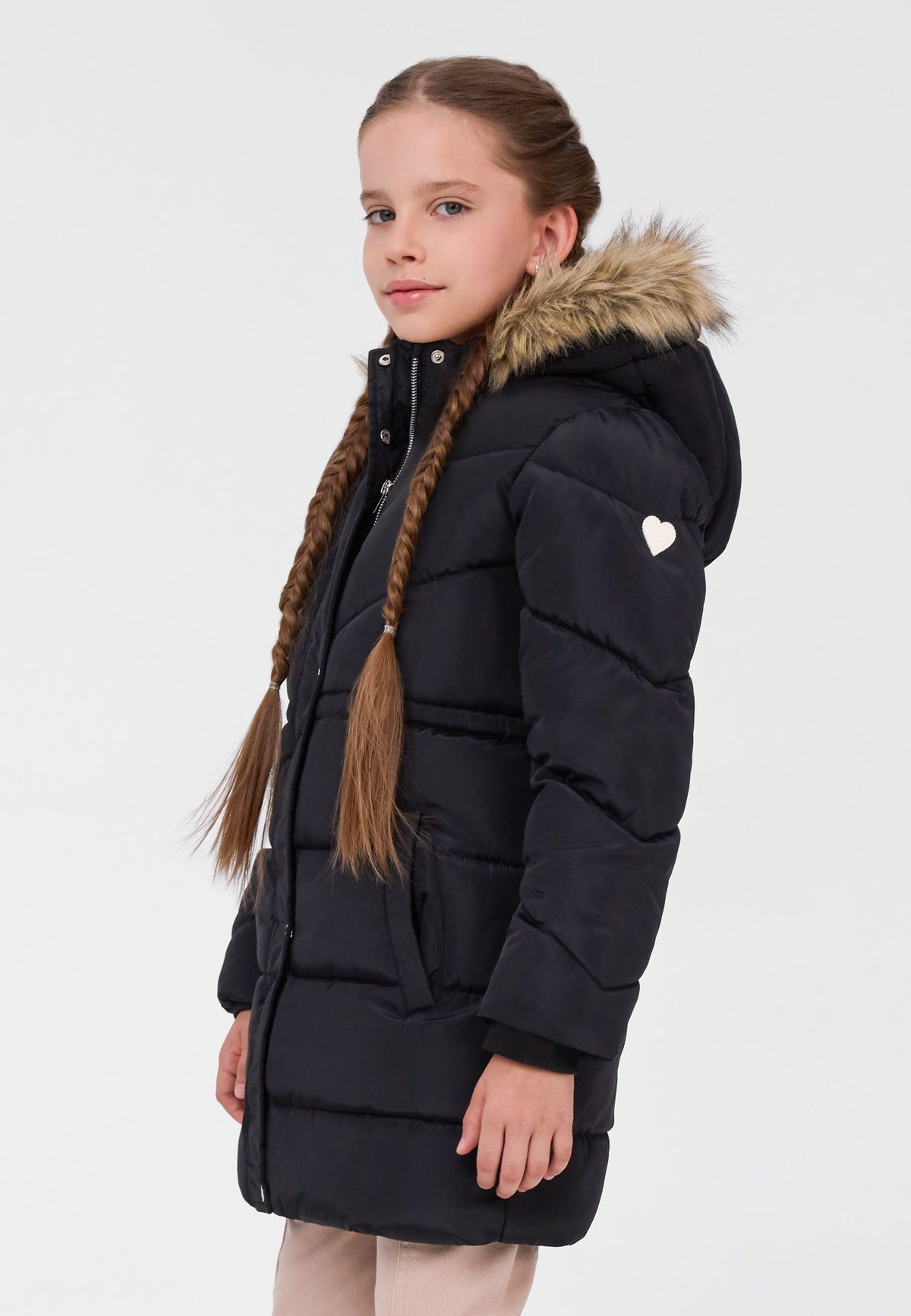 Girl in side view wearing a long black padded coat with beige faux-fur hood trim and white heart on the sleeve.