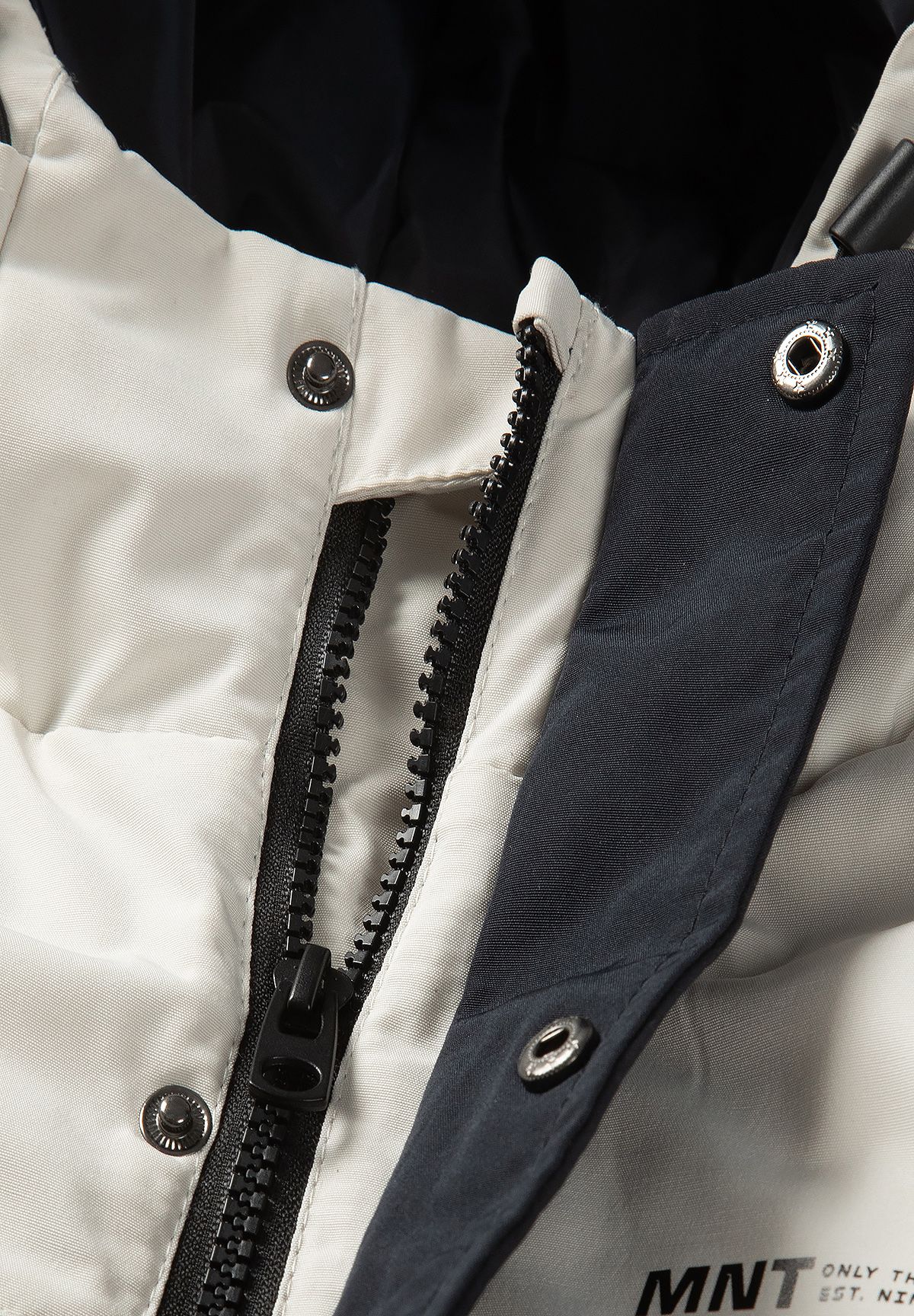 Close-up of the parka's black centre zip beside a black storm flap with metal snaps on cream fabric.