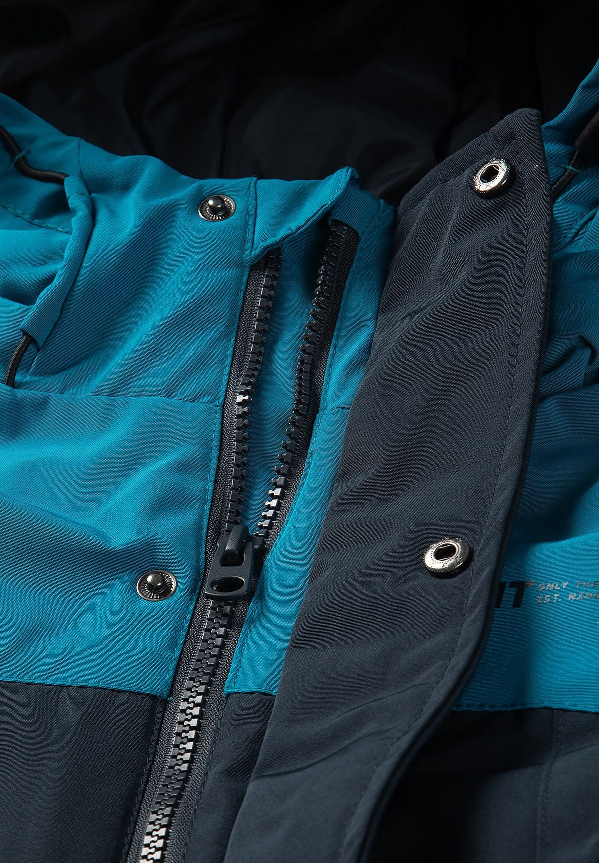 Close-up of the parka's black centre zip beside a navy storm flap with metal snaps on teal fabric.