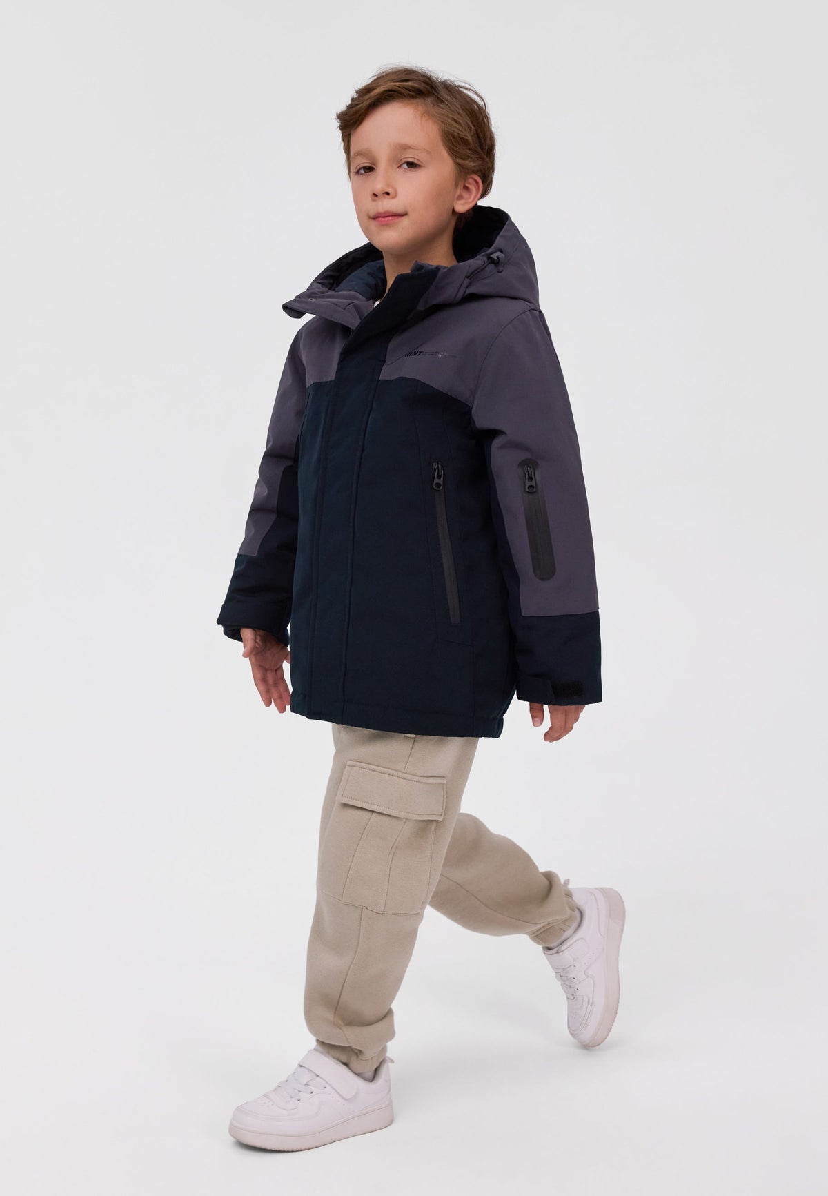 Boy wearing a charcoal and black hooded parka with a zipped sleeve pocket and long front pocket zips.