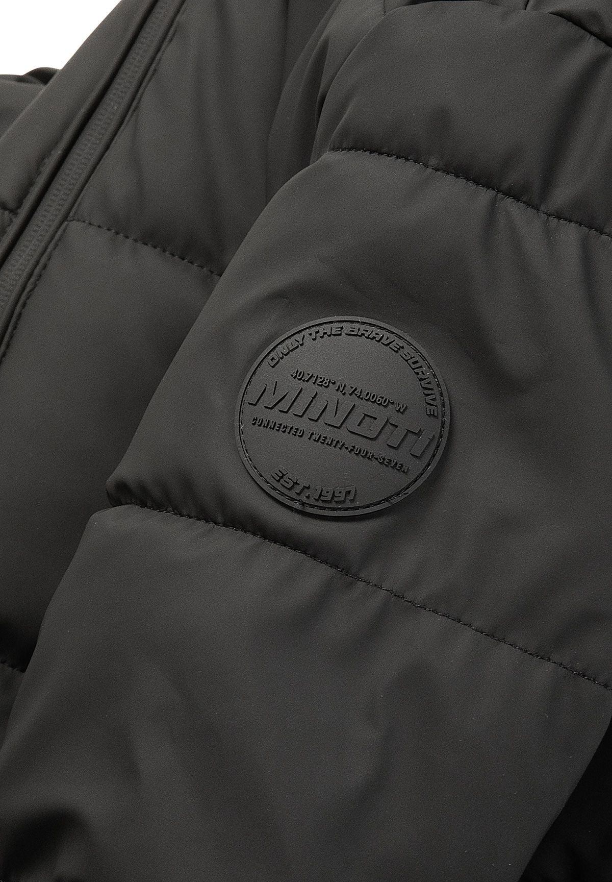 Close-up of a circular black Minoti emblem embossed on the matte black puffer sleeve.