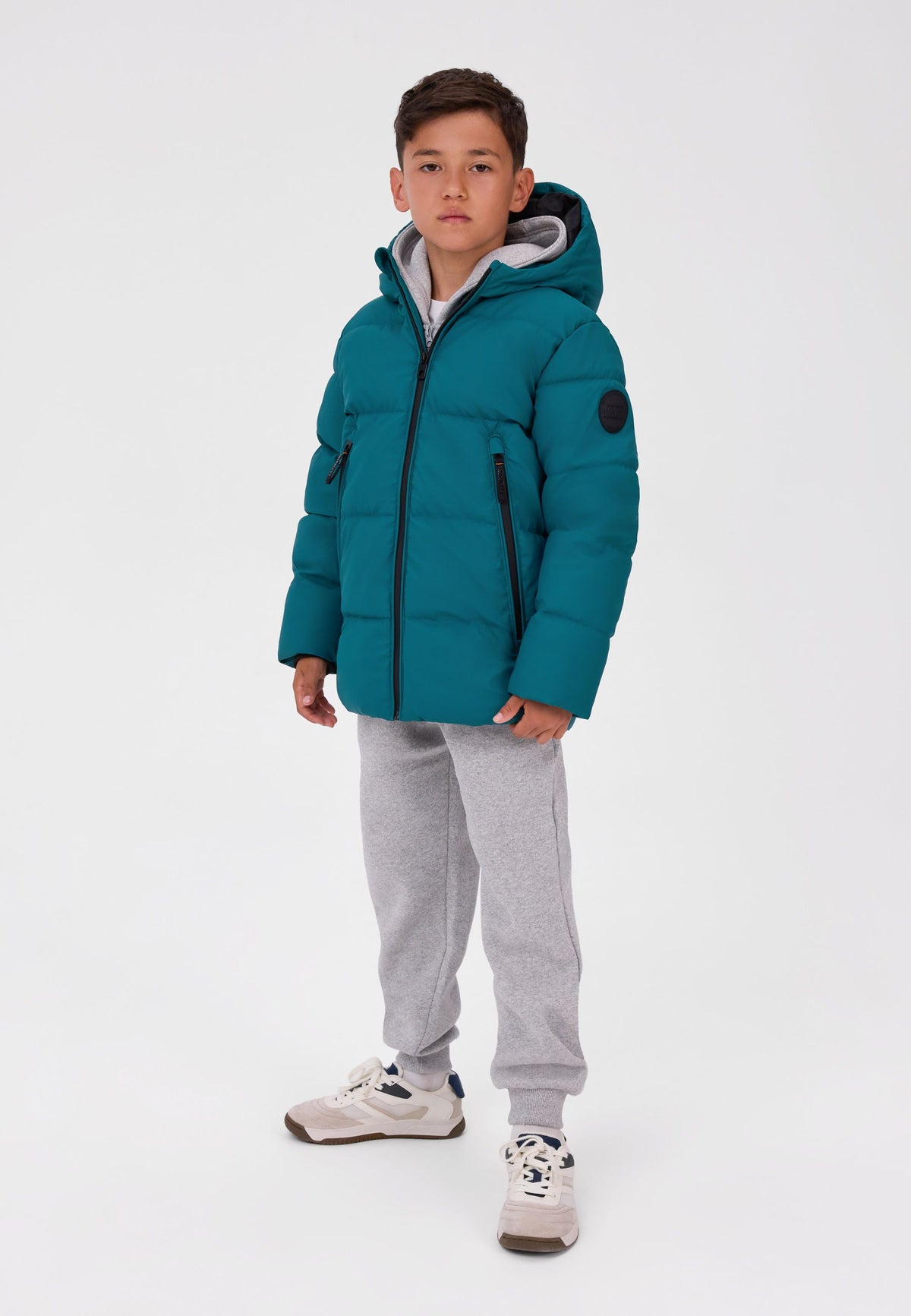 Boy wearing a teal hooded puffer jacket with black zip, two zipped pockets and round sleeve badge.