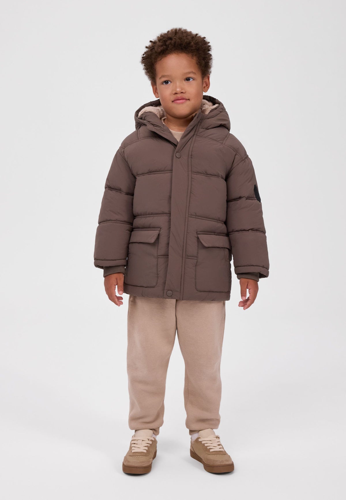 Boy wearing a brown hooded padded jacket with two lower flap pockets and beige teddy lining.