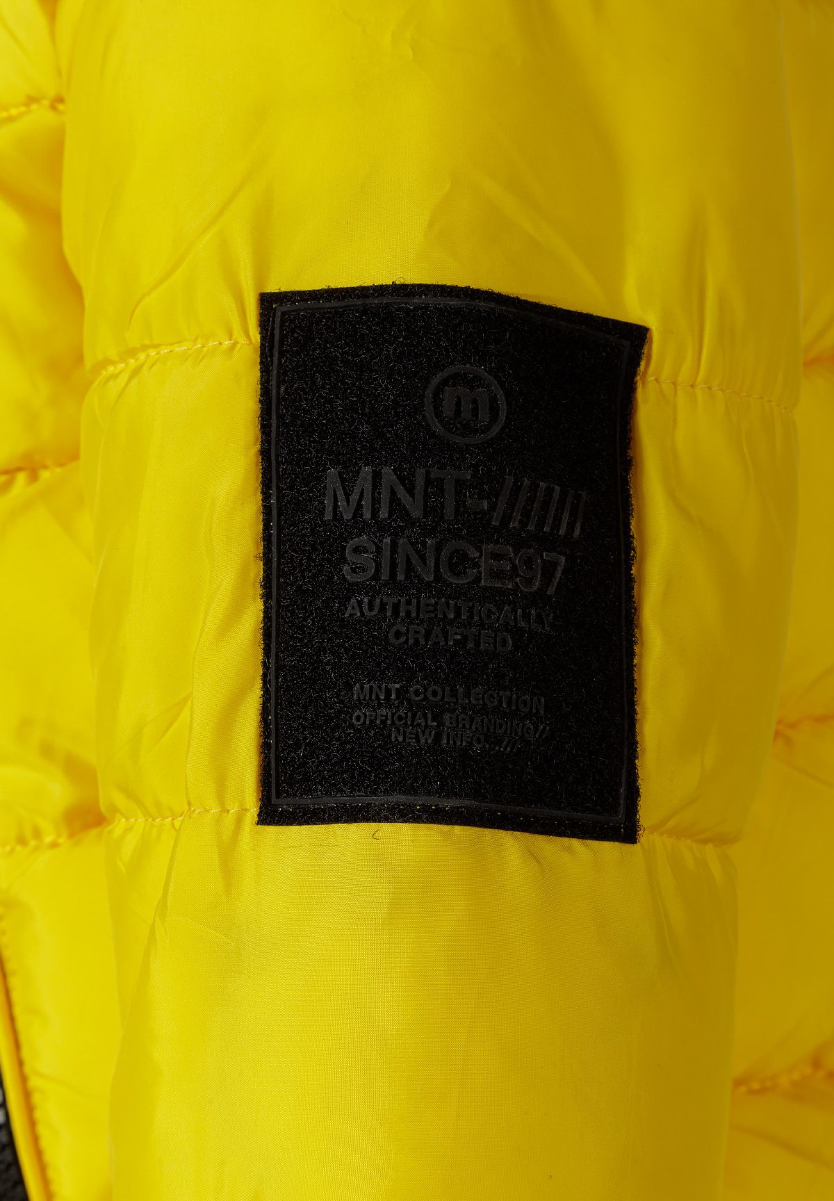 Close-up of a black MNT Since 97 rectangular patch on the yellow jacket sleeve.