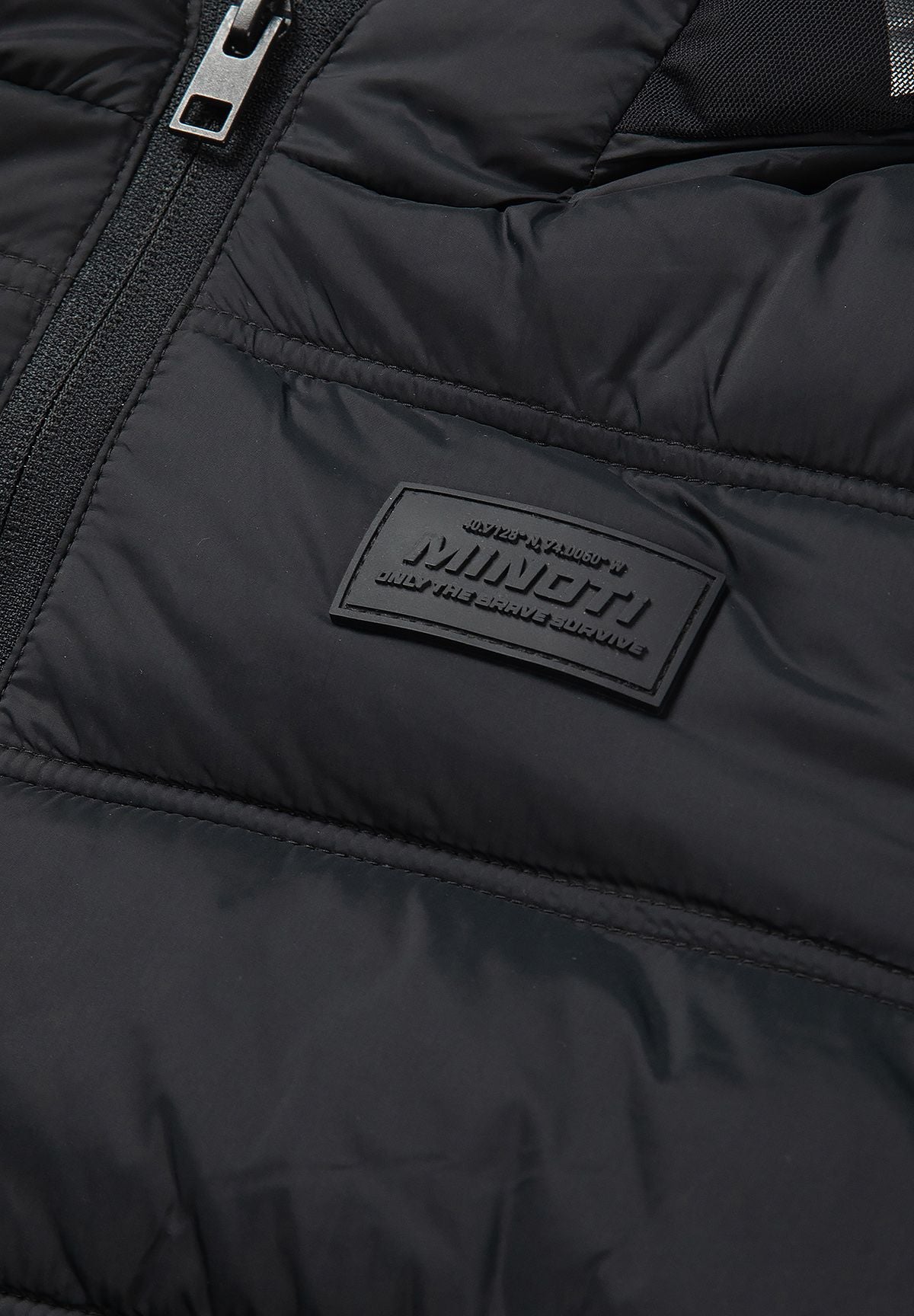 Close-up of the tonal rectangular MINOTI badge on the black jacket's quilted chest.