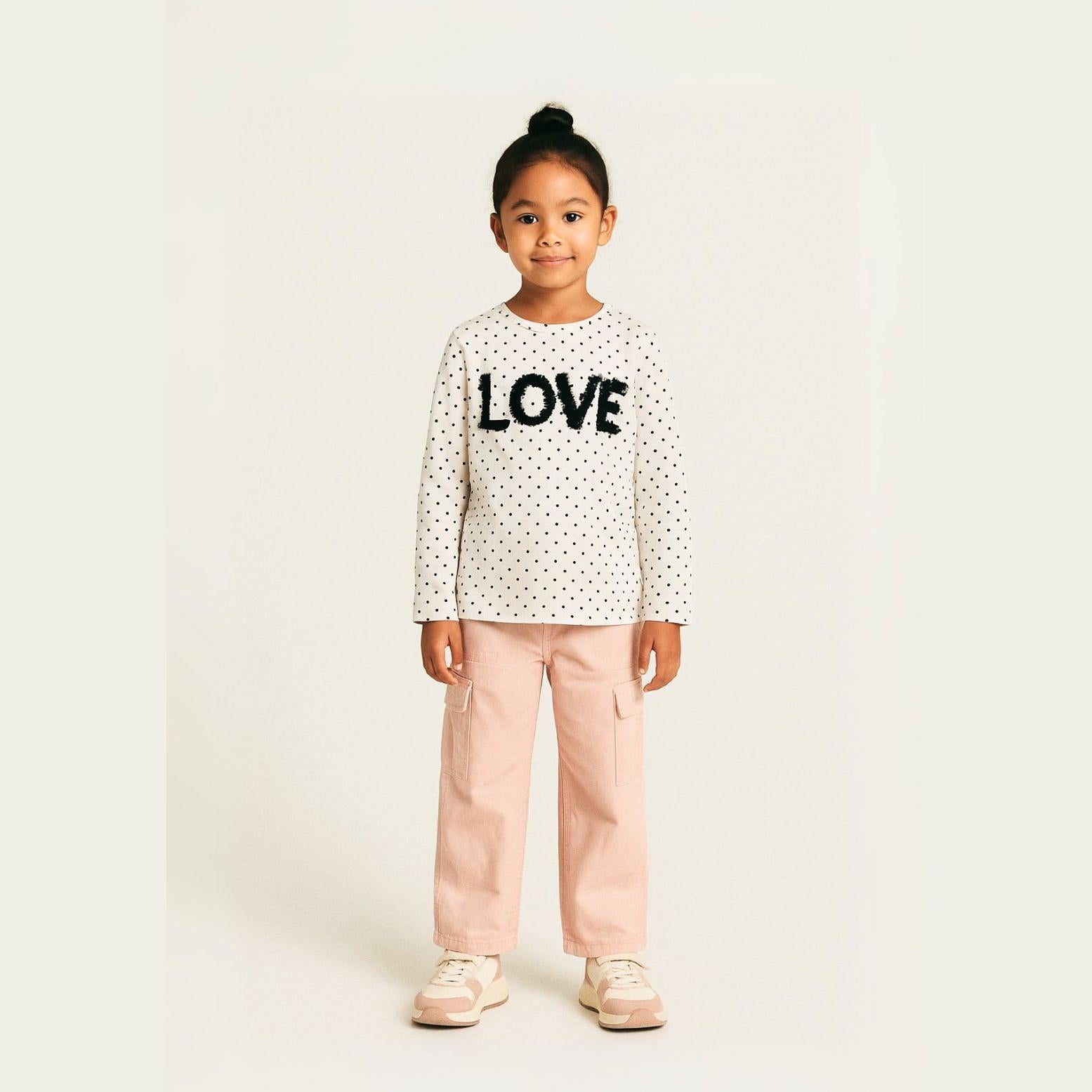 Young girl wearing white polka dot long sleeve top with LOVE applique and pink trousers, standing against neutral background