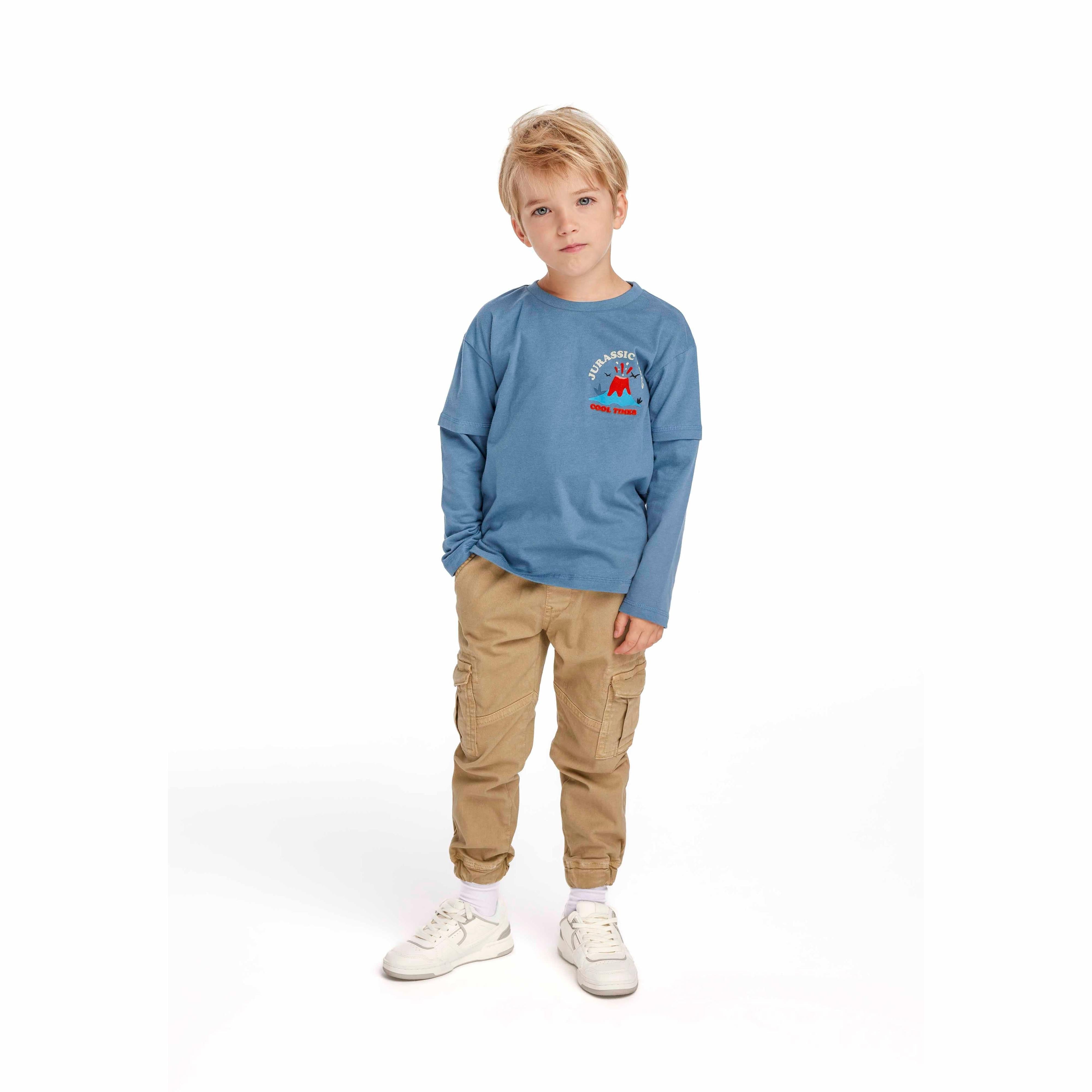 Young blonde boy wearing navy long sleeve t-shirt with Jurassic Vibes volcano print and beige cargo pants, standing against white background