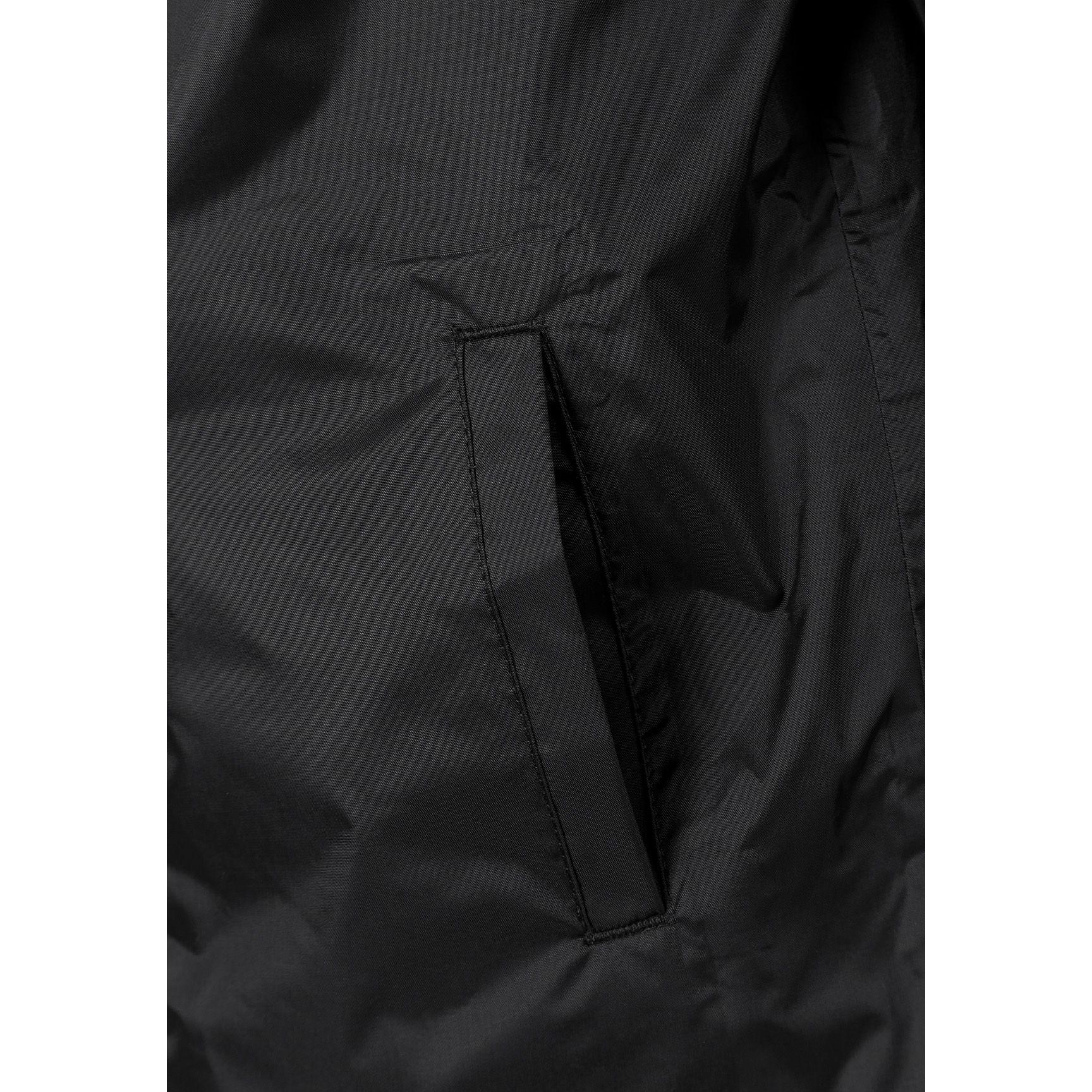 Boy in black hooded waterproof jacket pointing forward, wearing light blue cargo jeans and black sneakers on white studio background