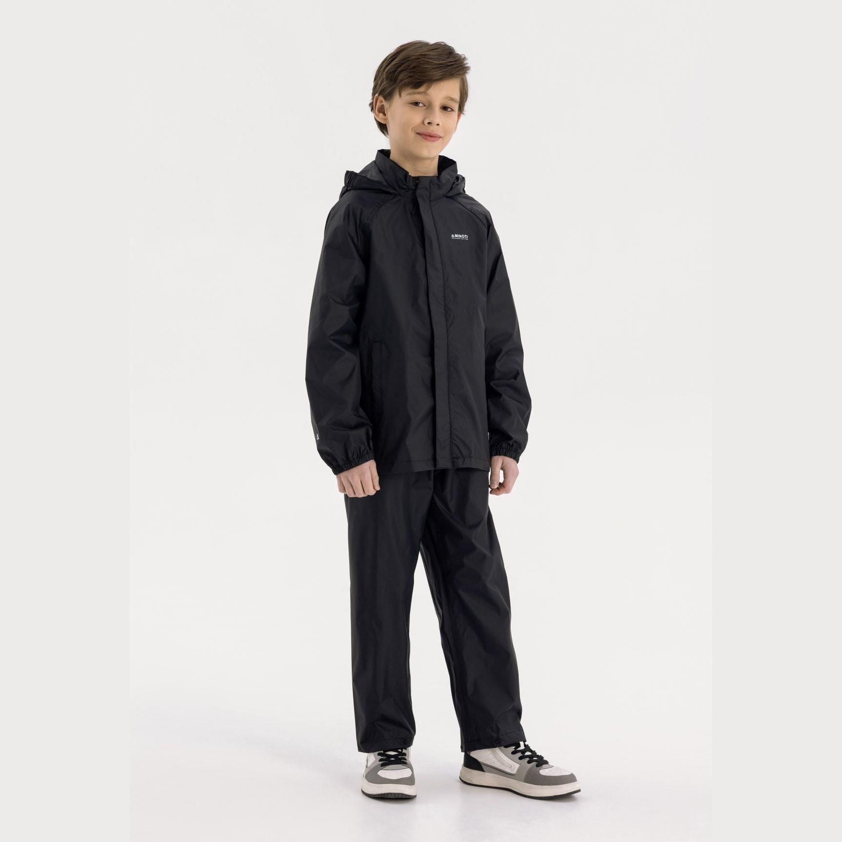 Boy in black hooded waterproof jacket pointing forward, wearing light blue cargo jeans and black sneakers on white studio background