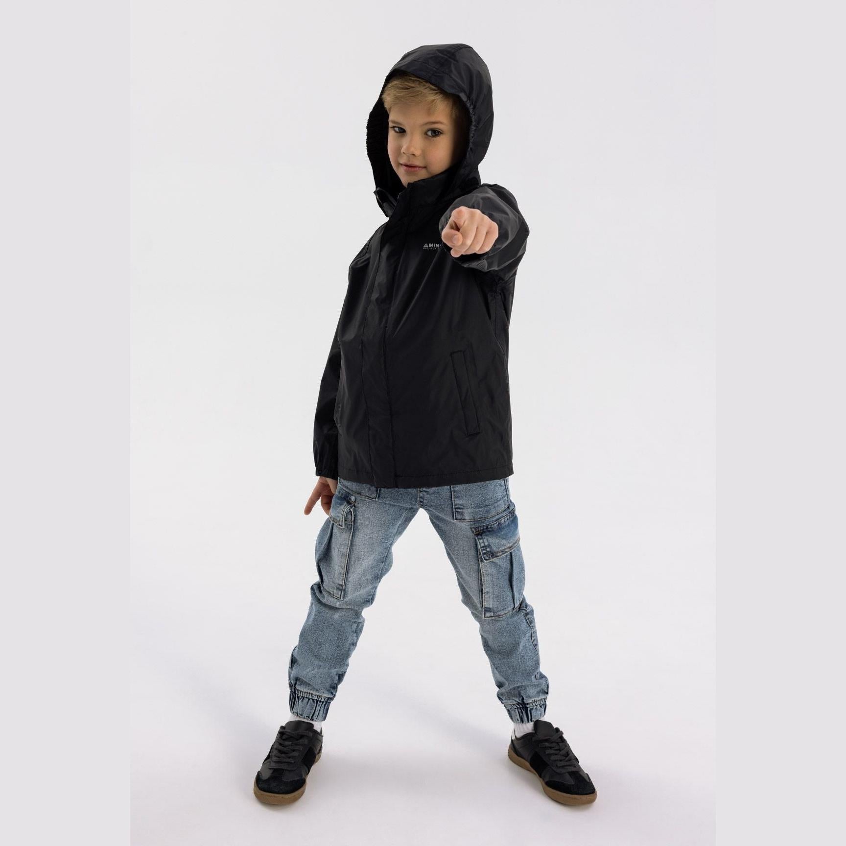 Boy in black hooded waterproof jacket pointing forward, wearing light blue cargo jeans and black sneakers on white studio background