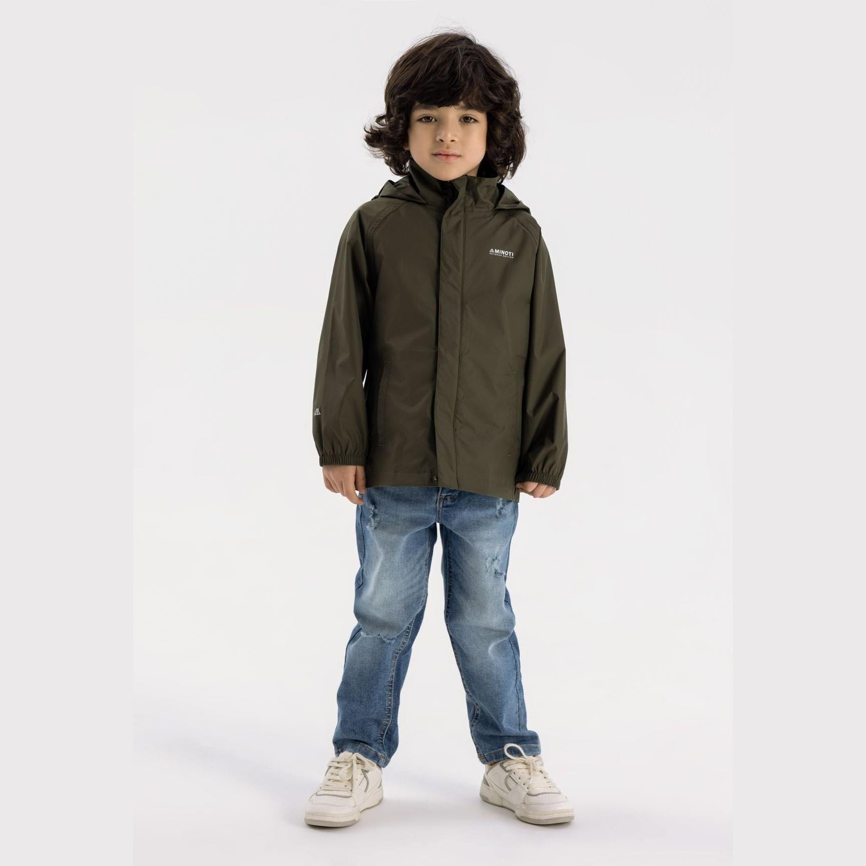 Young boy wearing khaki hooded waterproof jacket with jeans and sneakers, standing against white background