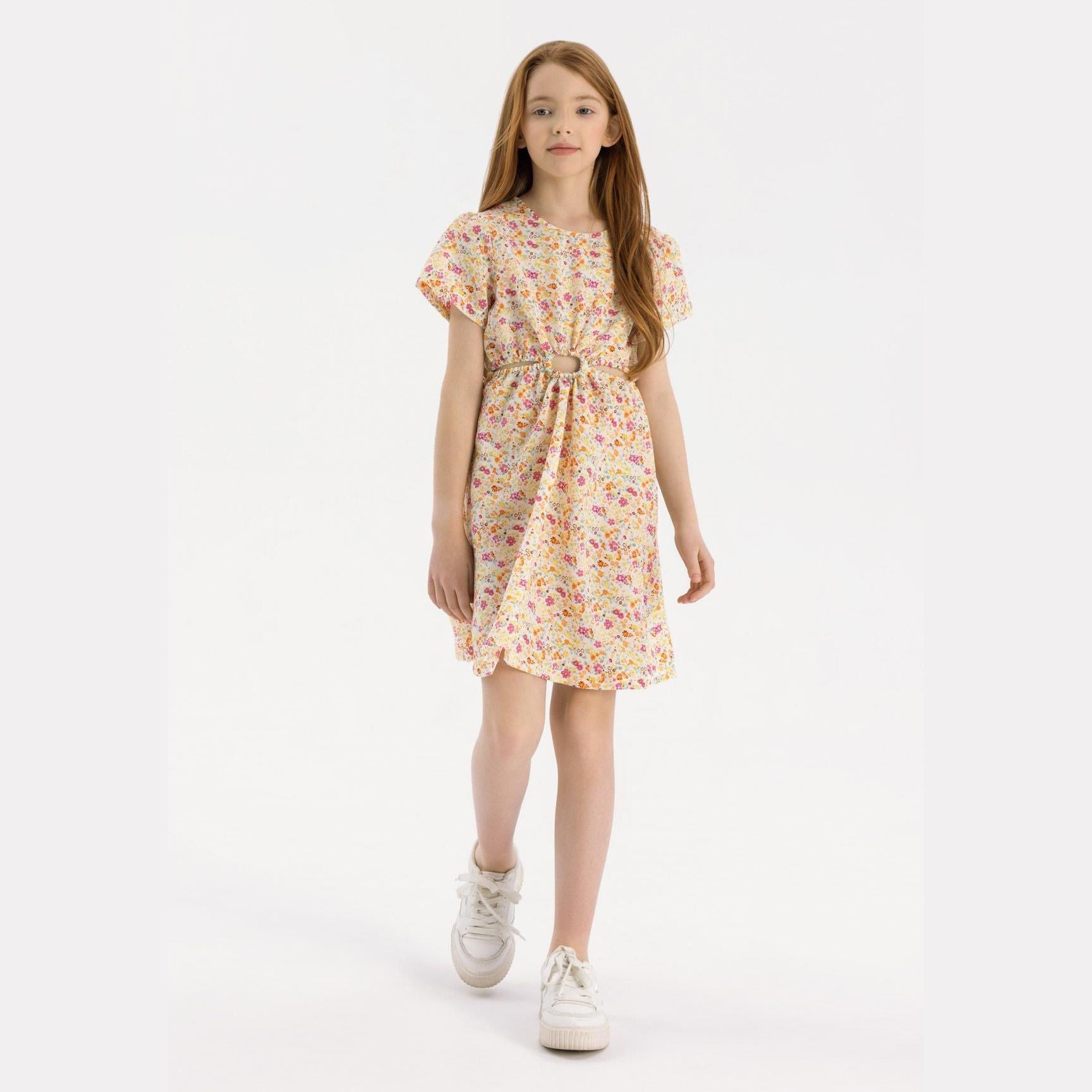 Young girl wearing yellow floral crinkle dress with cut-out waist and ruffle sleeves, standing against white background wearing white sneakers