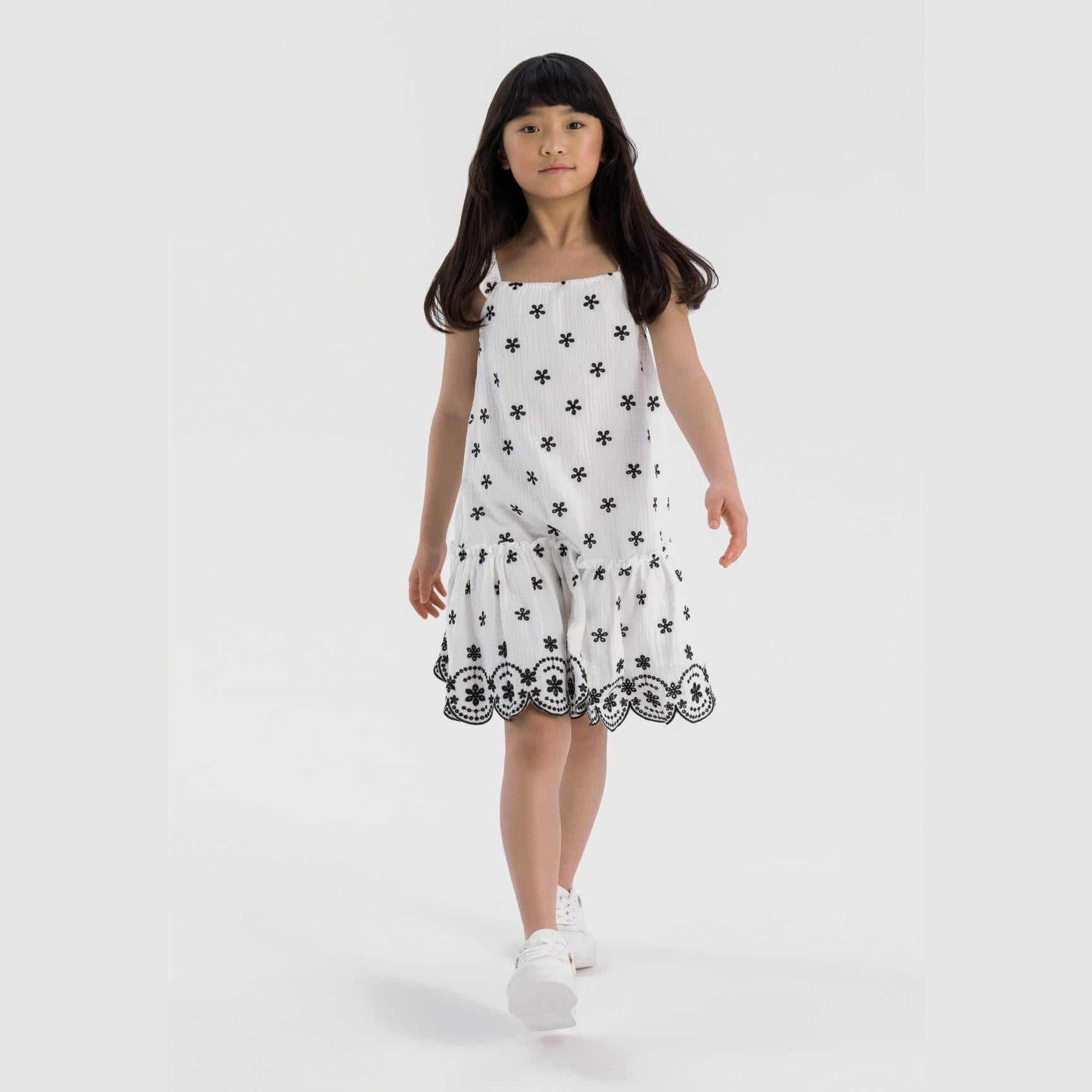 Young girl wearing white cotton muslin dress with black floral embroidery and scalloped hem, standing against light grey background