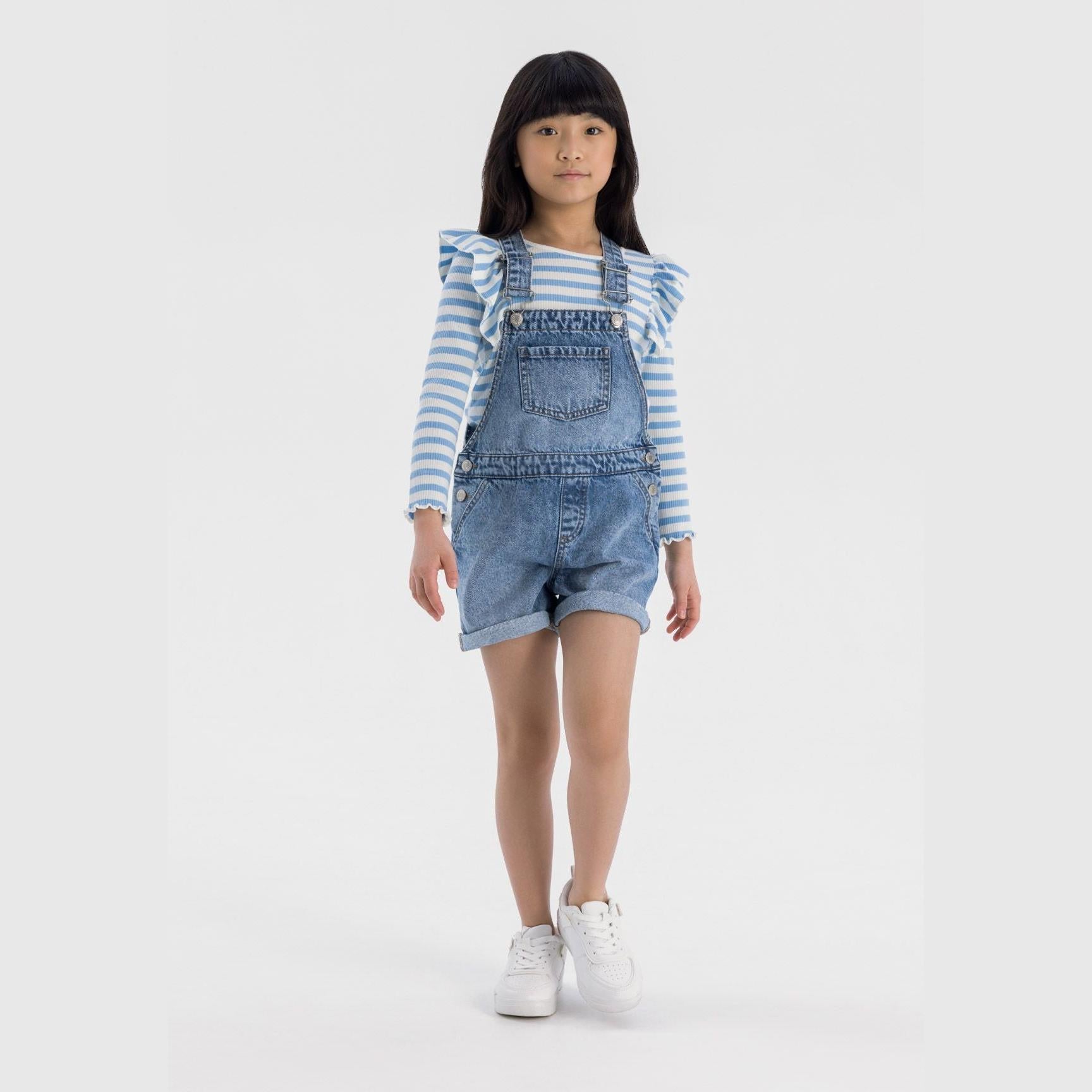 Young girl wearing navy denim short dungarees with blue and white striped long-sleeve top, standing against white studio background