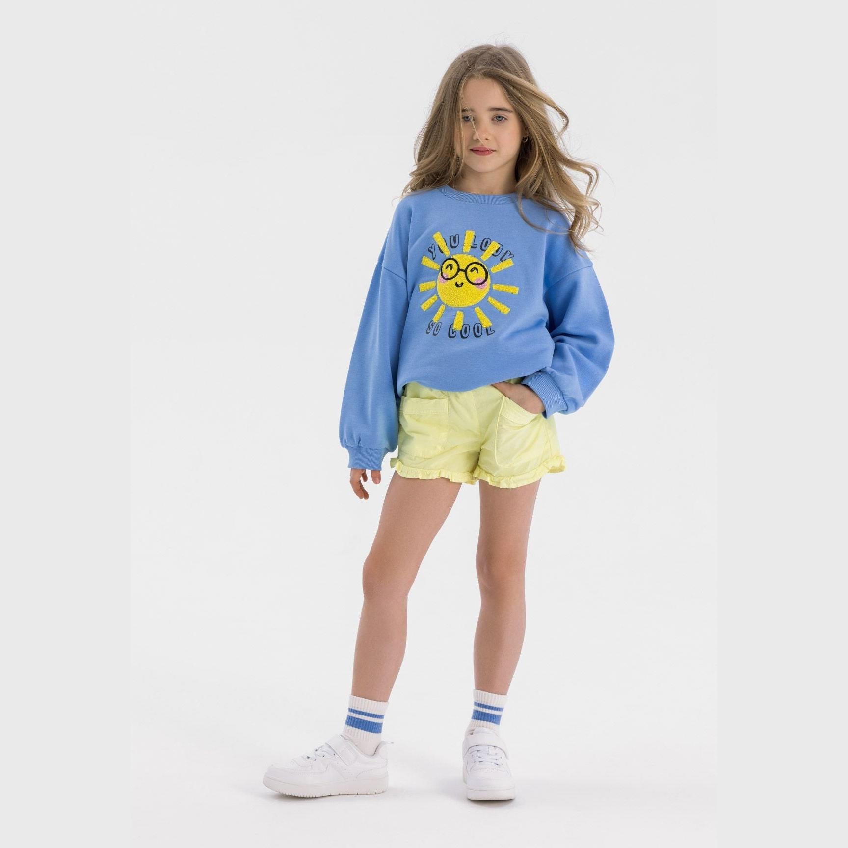 Young girl wearing light blue fleece sweatshirt with yellow sun print, paired with yellow shorts and white sneakers, standing against white background