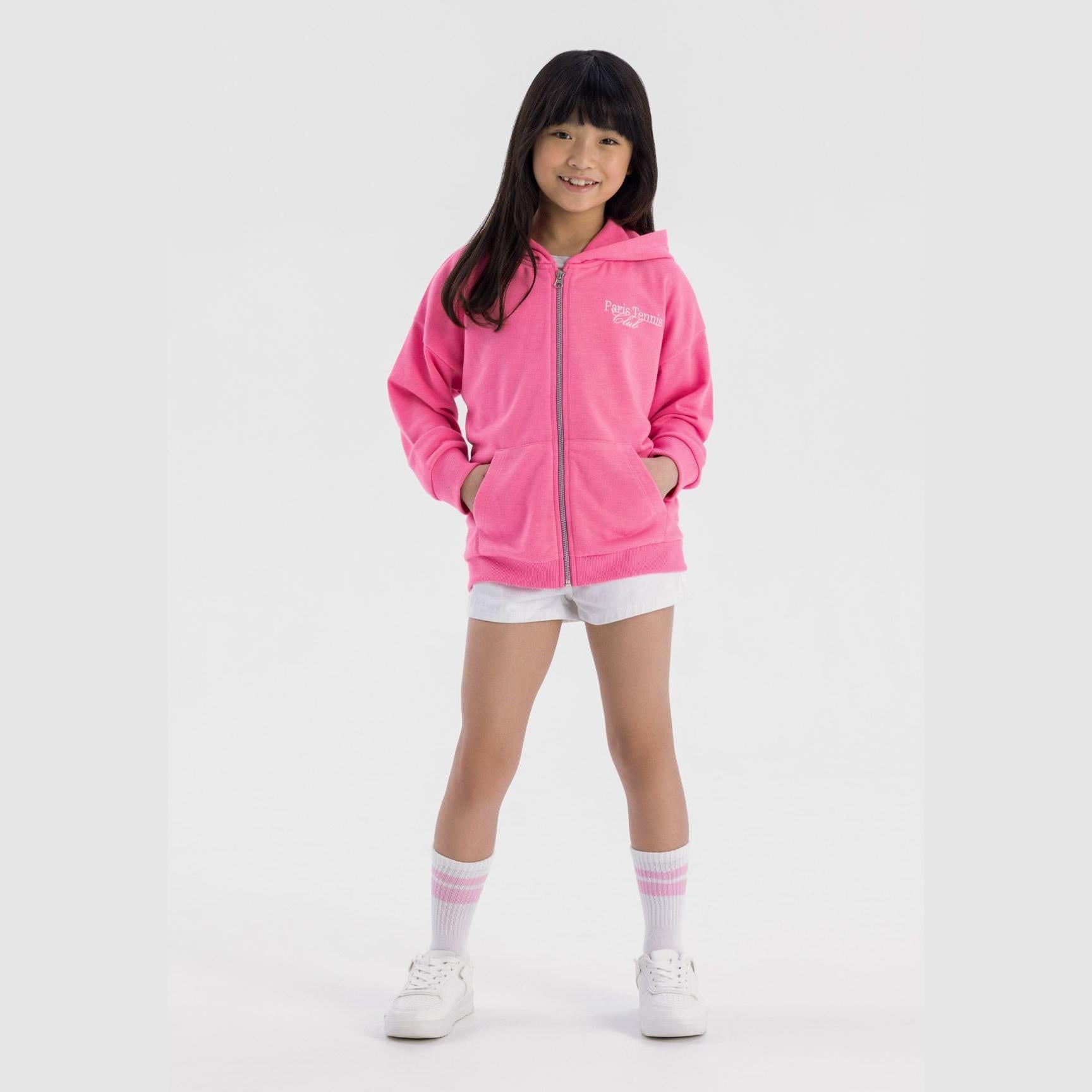 Young girl wearing oversized pink zip hoodie with Paris Tennis Club embroidery, paired with white shorts and striped socks, standing against white background