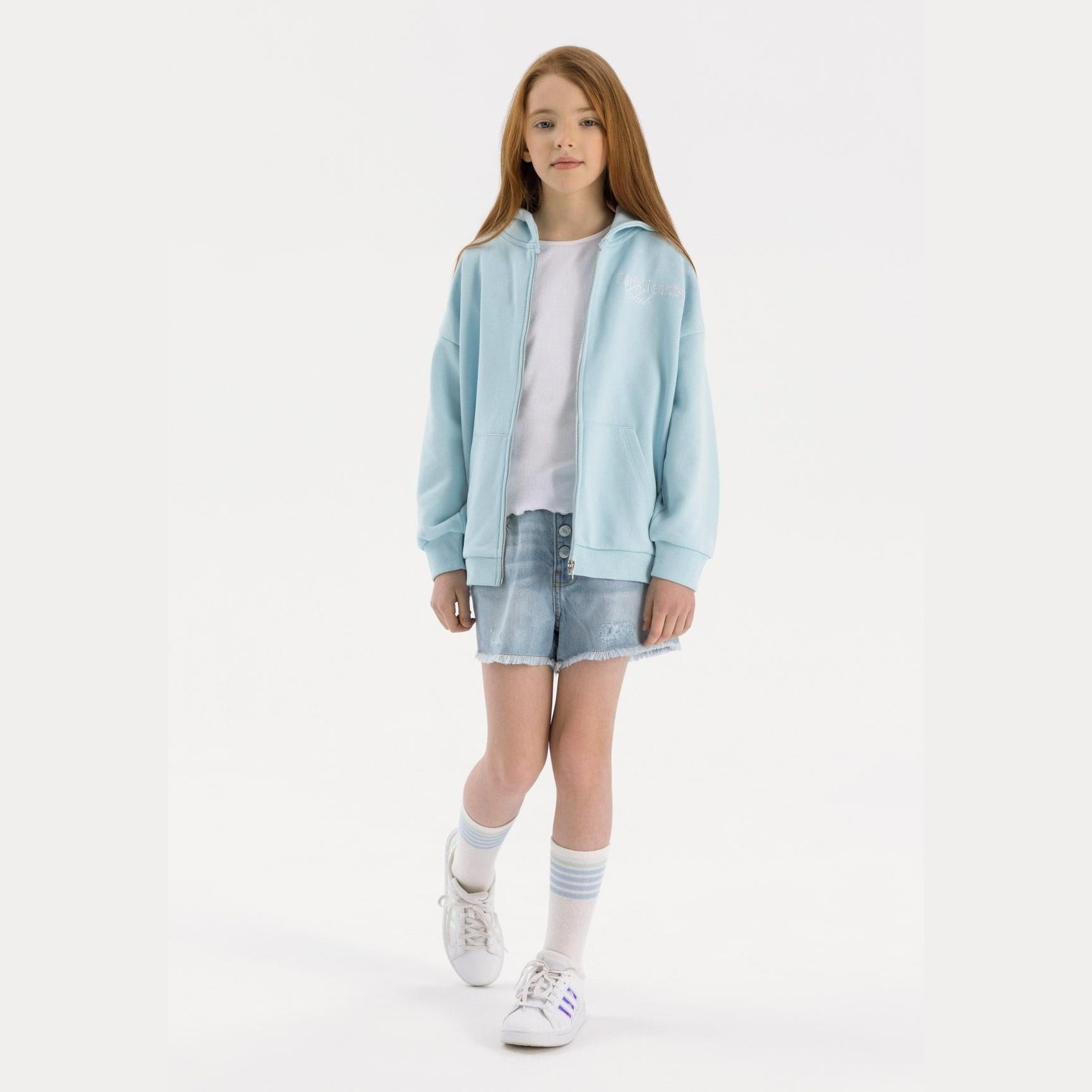 Young girl wearing light blue oversized zip hoodie with white t-shirt, denim shorts and white sneakers, standing against white background